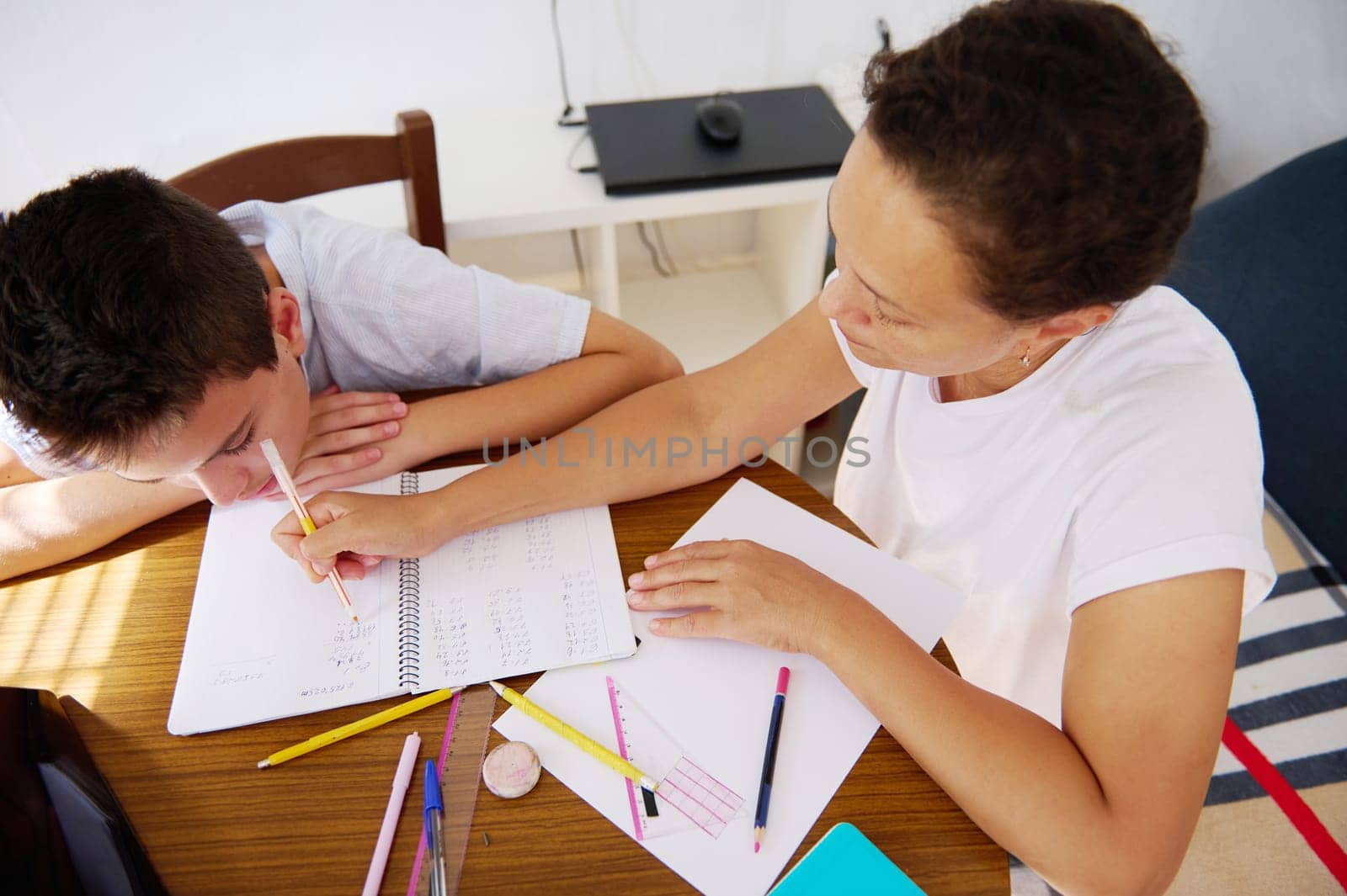 A mother assists her son with his homework at the kitchen table. Educational materials, notebooks, and pencils are spread out, emphasizing learning and support in a home setting.