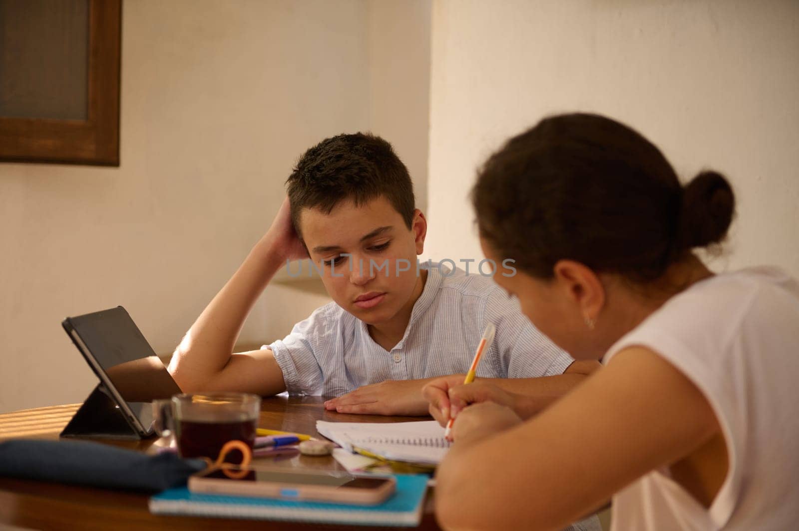 A mother and her son are focused on studying together at a table. The son is using a tablet while the mom assists with his homework, creating a supportive learning environment.