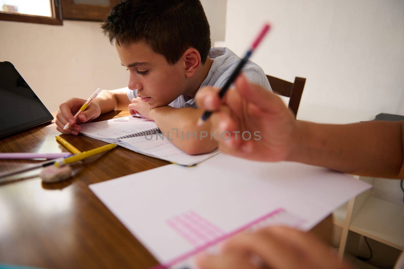 A young boy concentrates on his homework at a wooden table, surrounded by school supplies and a digital tablet, emphasizing focus and education.