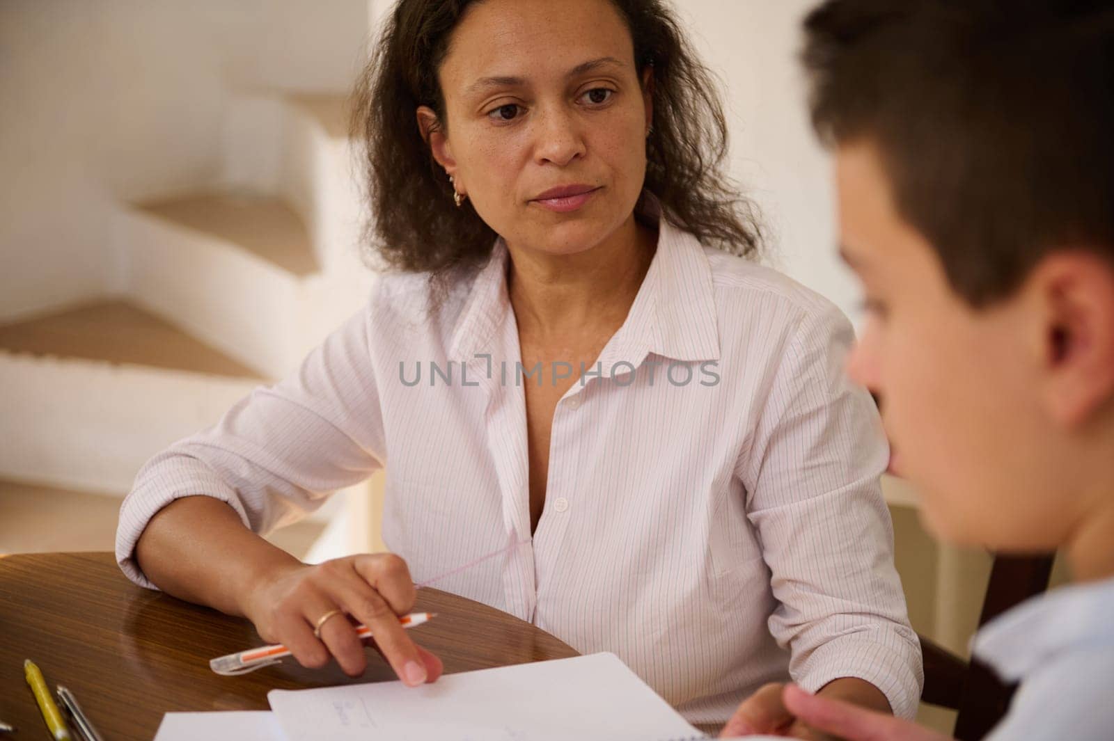 A mother attentively helps her son with homework at a wooden dining table. They are engaged in a focused conversation, highlighting the importance of education and parental involvement.