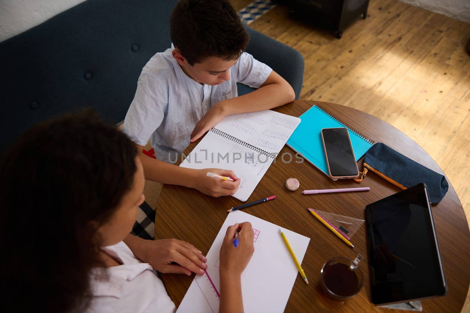 A mother and son are studying together at a wooden table, focusing on math problems. The scene includes notebooks, pencils, a tablet, and a cup of tea, creating a productive learning environment.