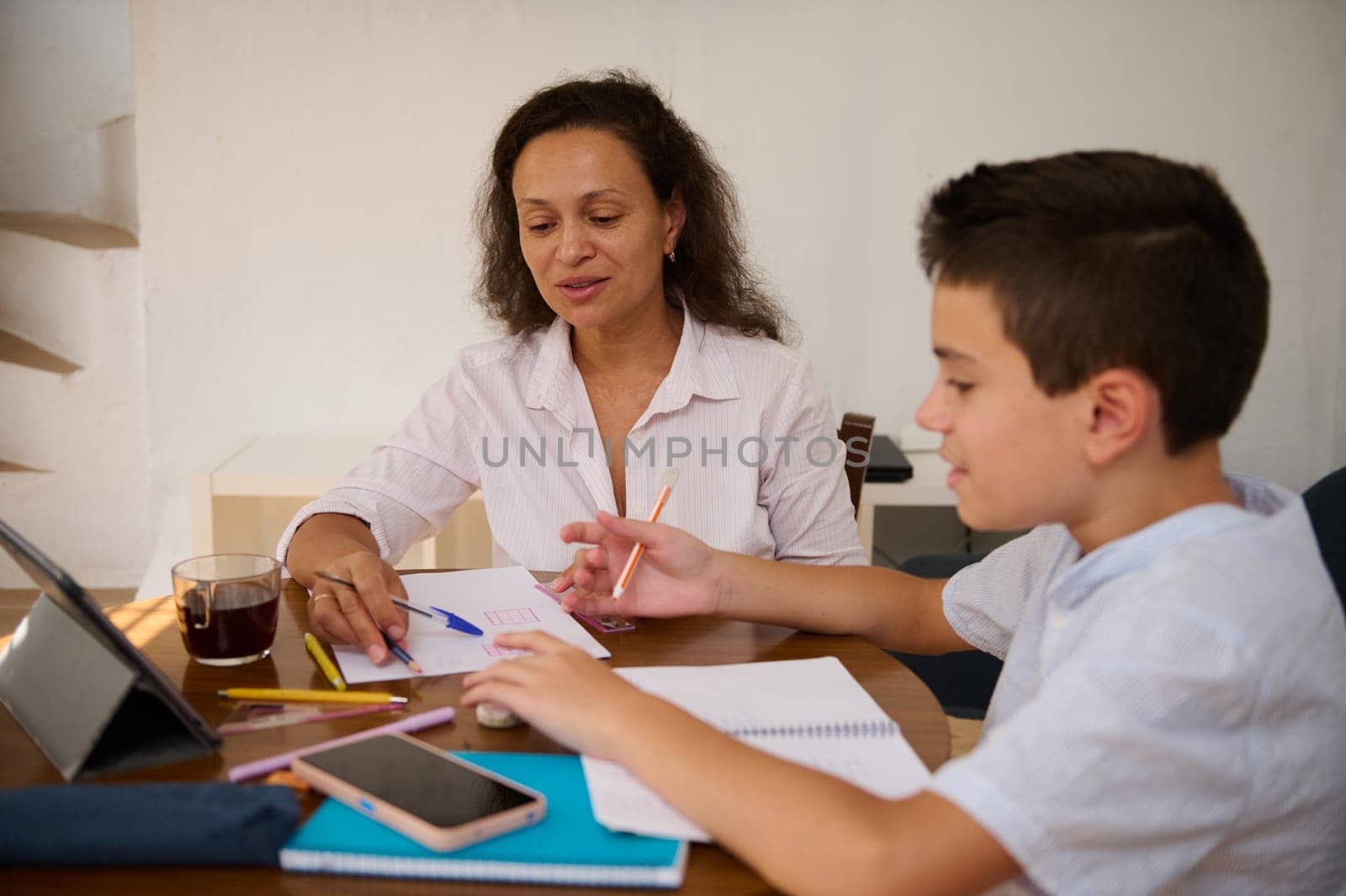 A mother assists her son with homework while sitting at a table. They are using a tablet and surrounded by notebooks and pencils, fostering a supportive learning environment.