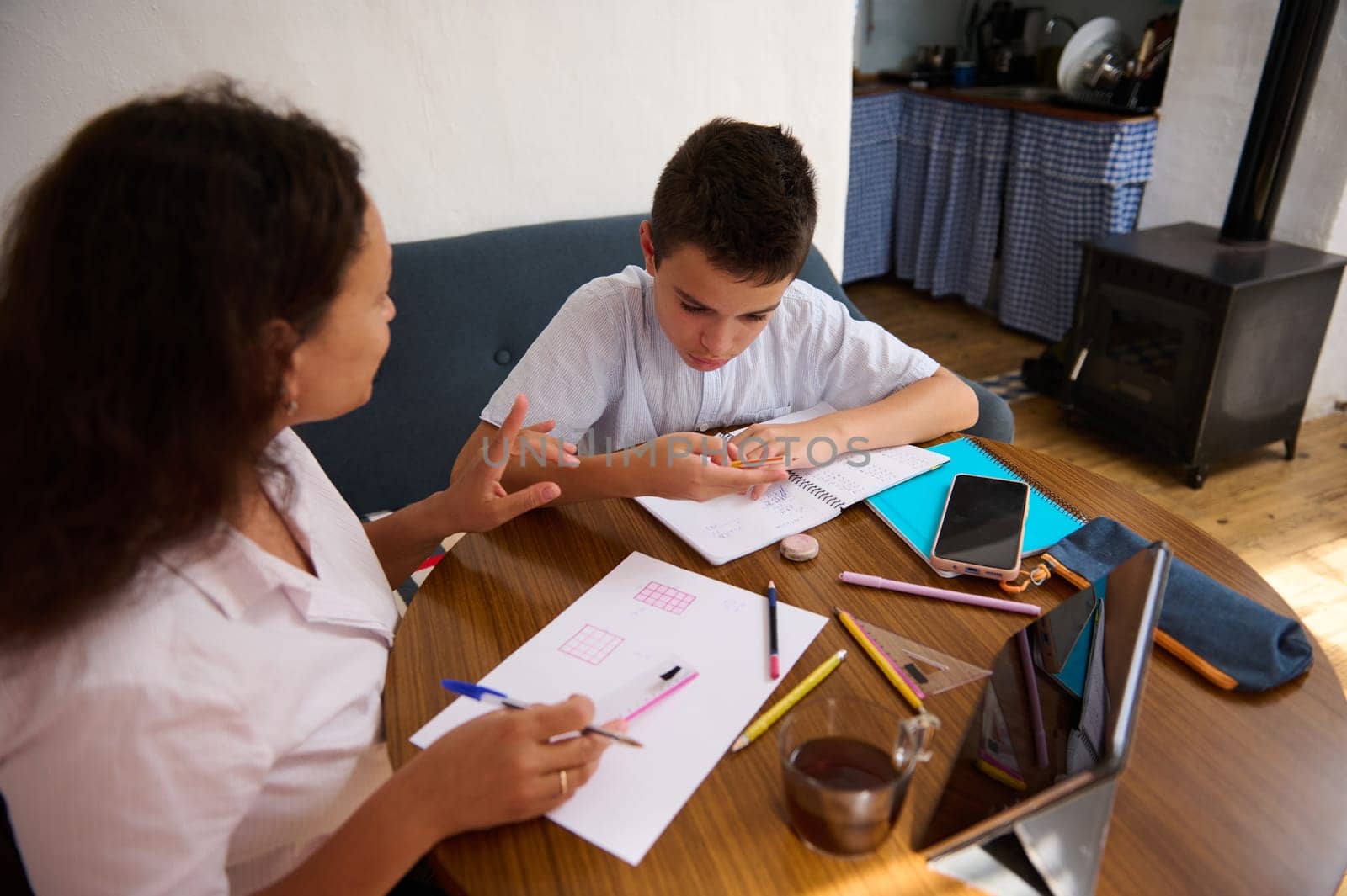 A mother helps her son with homework at the kitchen table, emphasizing concentration and learning. Books, tablets, and notebooks scattered around.