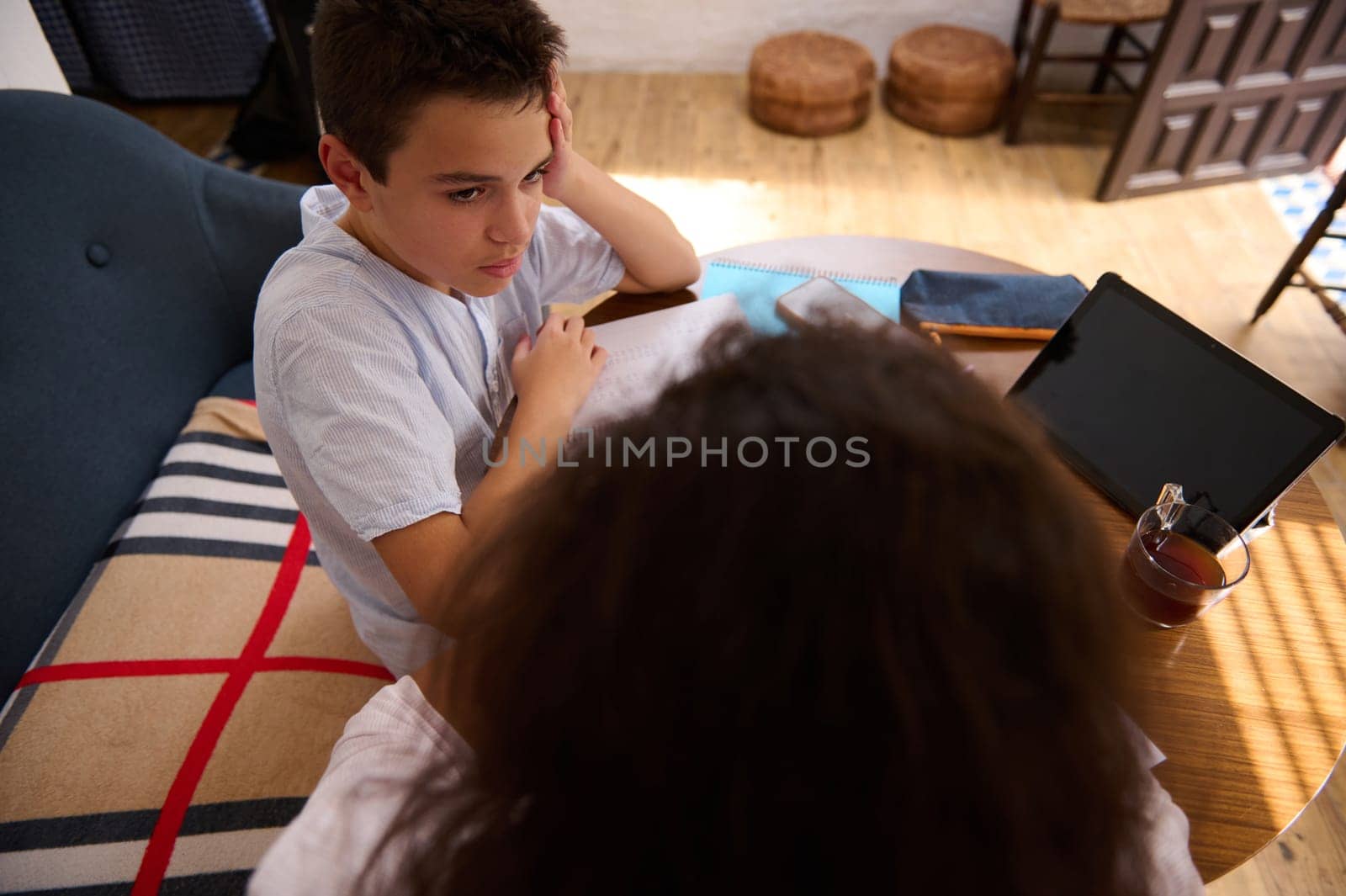 A young boy intensely studies with the help of a digital tablet at home, fostering education and learning. The cozy setting includes a blanket and drink, adding comfort to his focused study session.