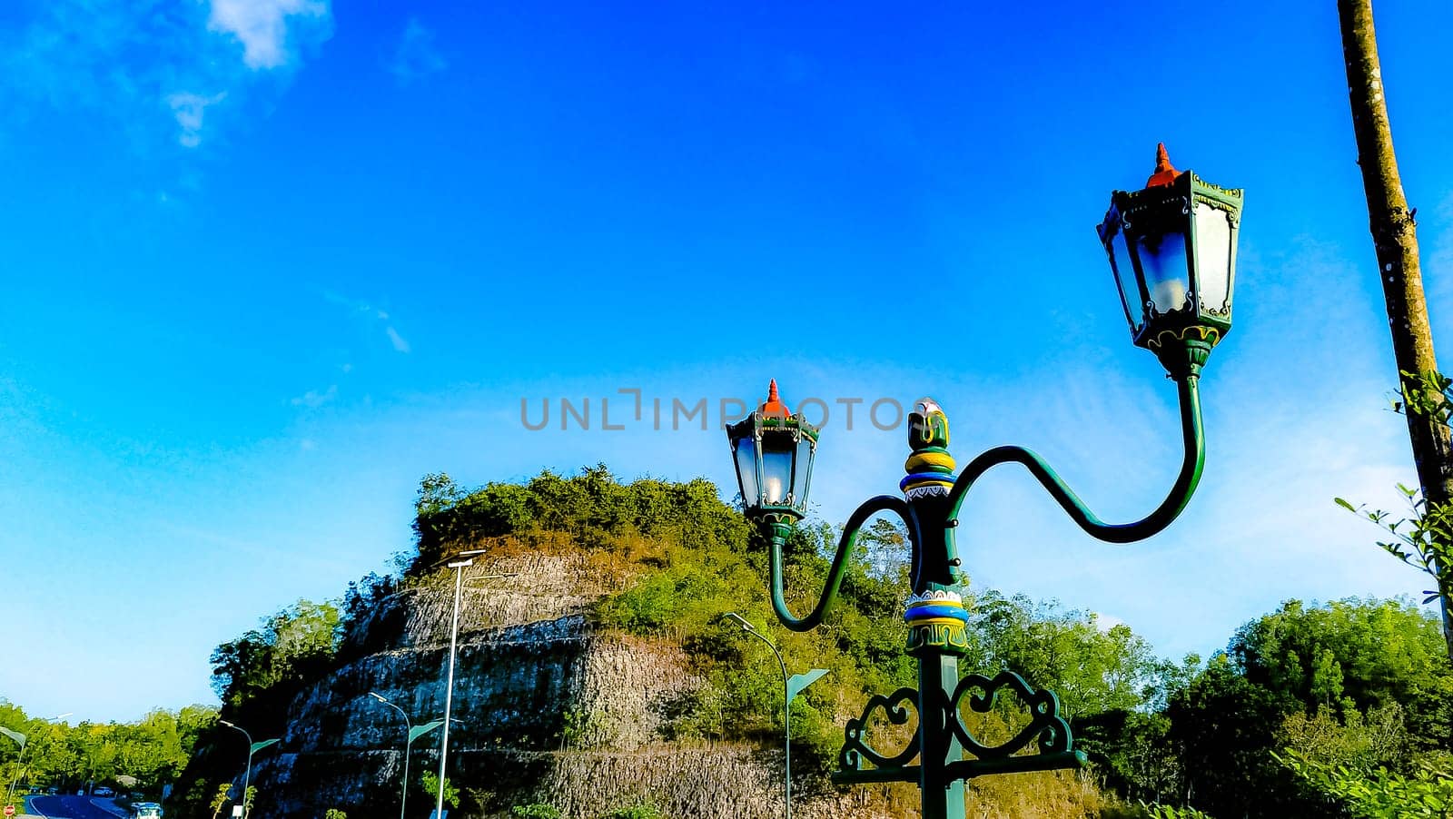 Elegant green street lamps with intricate designs stand in front of a vivid blue sky and lush hill. The scene exudes a blend of nature and historical architecture.