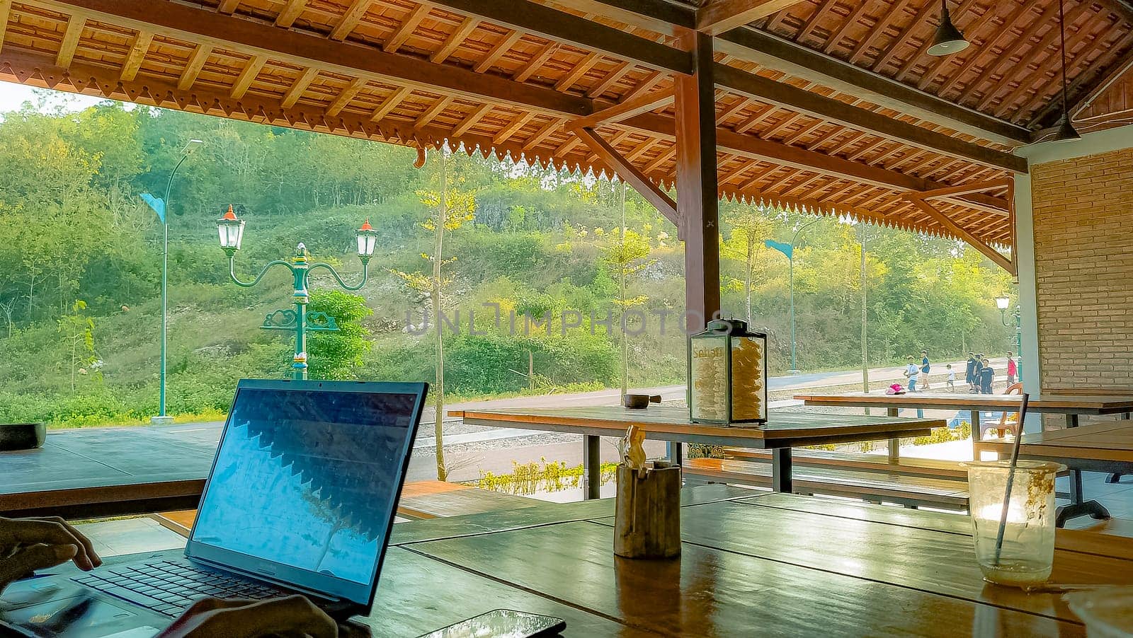 A serene outdoor workspace with a laptop on a wooden table, offering a peaceful view of nature and distant people enjoying the scenery.