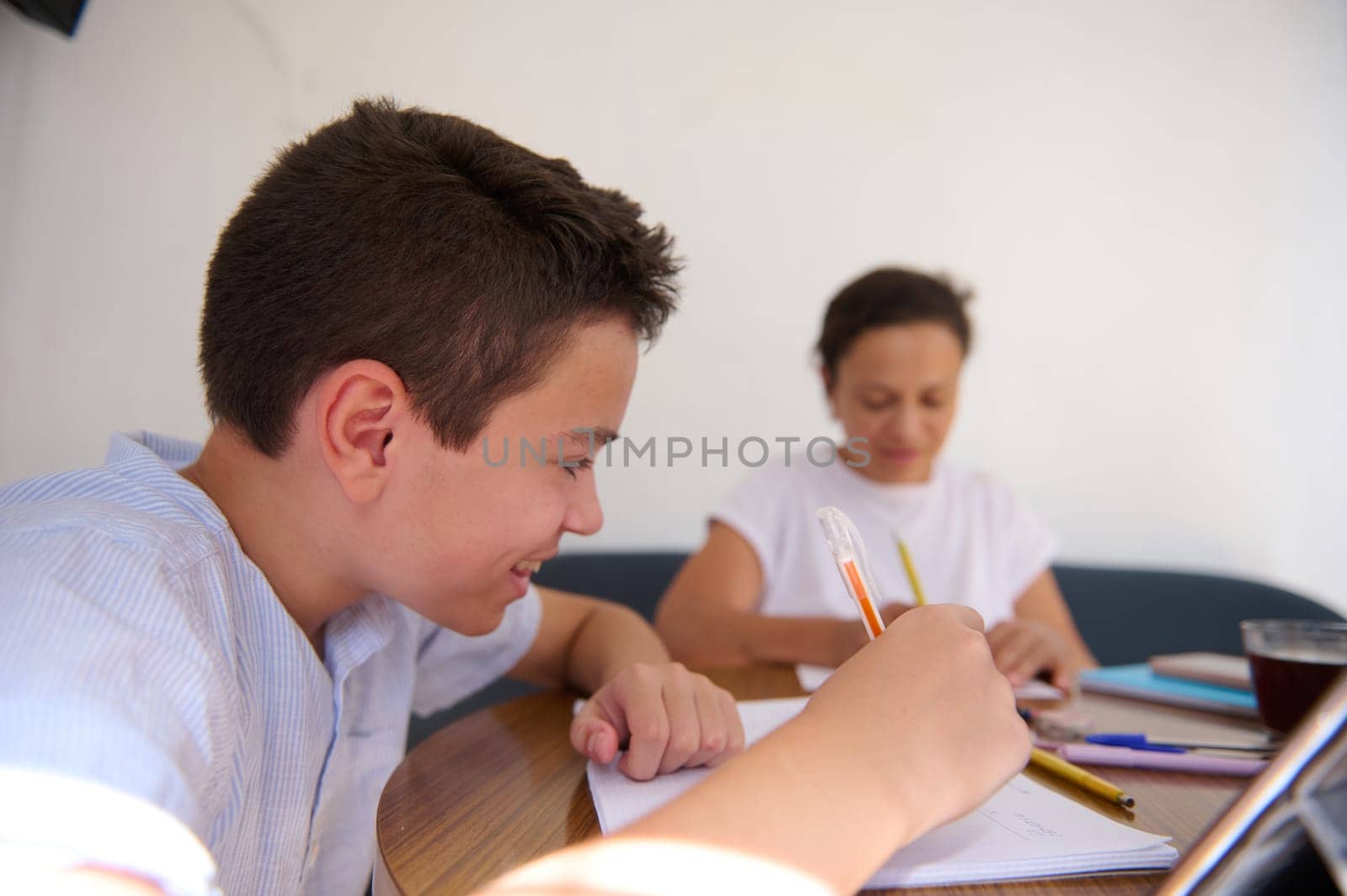 A mother and son work together at a table, focusing on homework with pens and paper in a bright setting, fostering learning and connection.