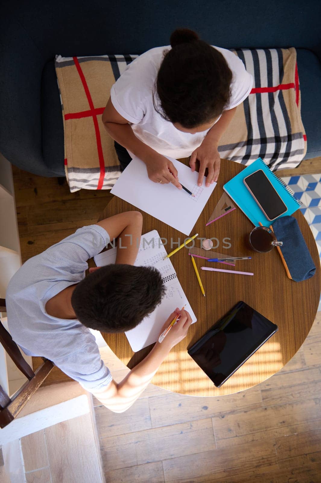 Two students working together at home on a project. Overhead view of a table with papers, tablets, and stationery, showing focused teamwork and learning.