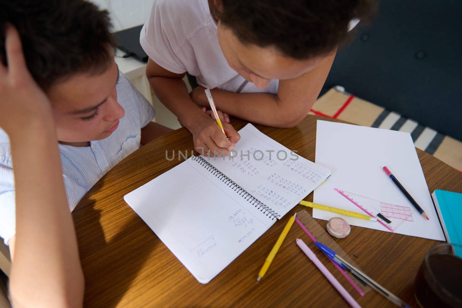 A mother assists her son with homework, focusing on mathematics. They sit at a kitchen table with notebooks and pens, sharing a moment of learning and encouragement.