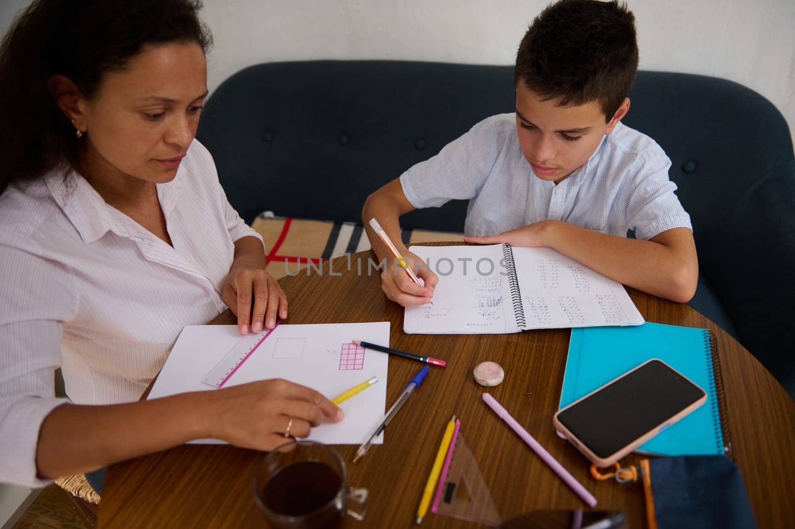 A mother and son sit together working on homework at a wooden table, surrounded by stationery. The mother guides her son with a ruler, depicting a supportive learning environment.