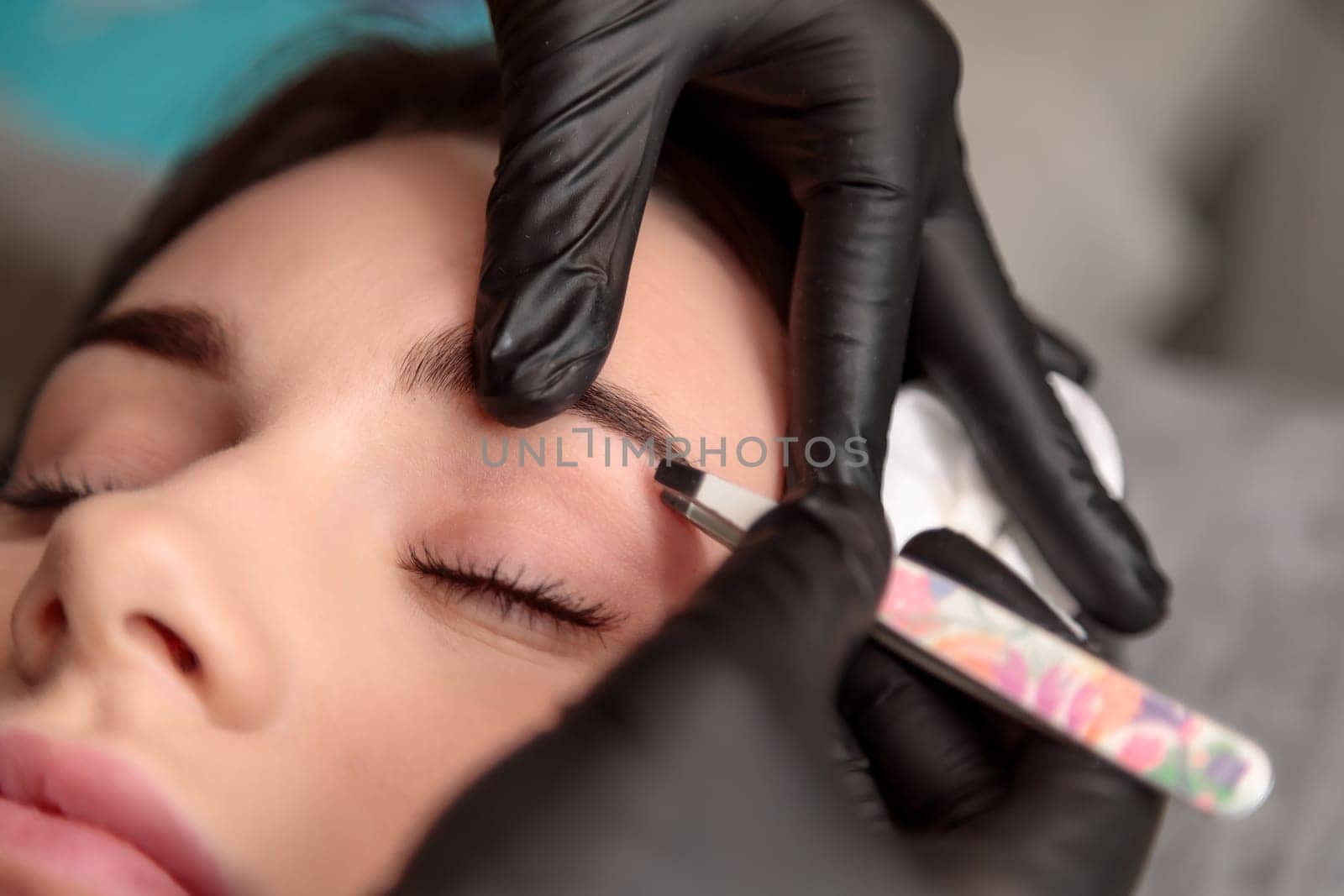 Beautician wearing black gloves using tweezers to shape eyebrows of client lying with closed eyes