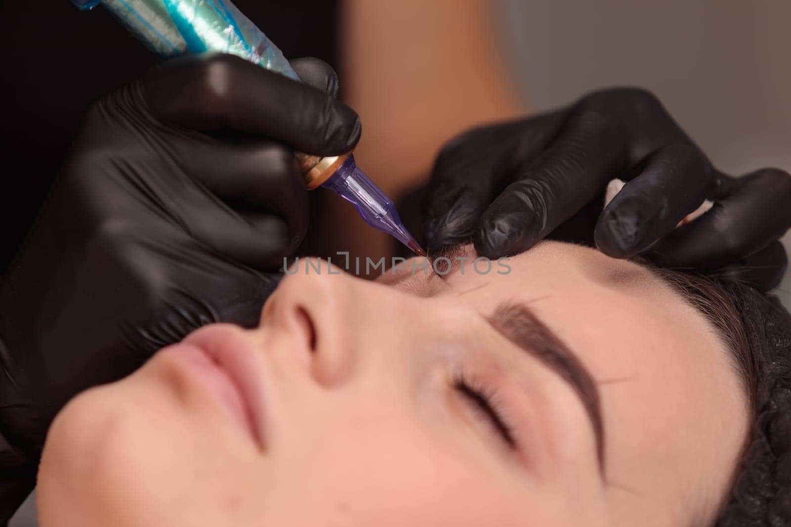 Cosmetologist making permanent eyebrow makeup using tattoo machine in beauty salon