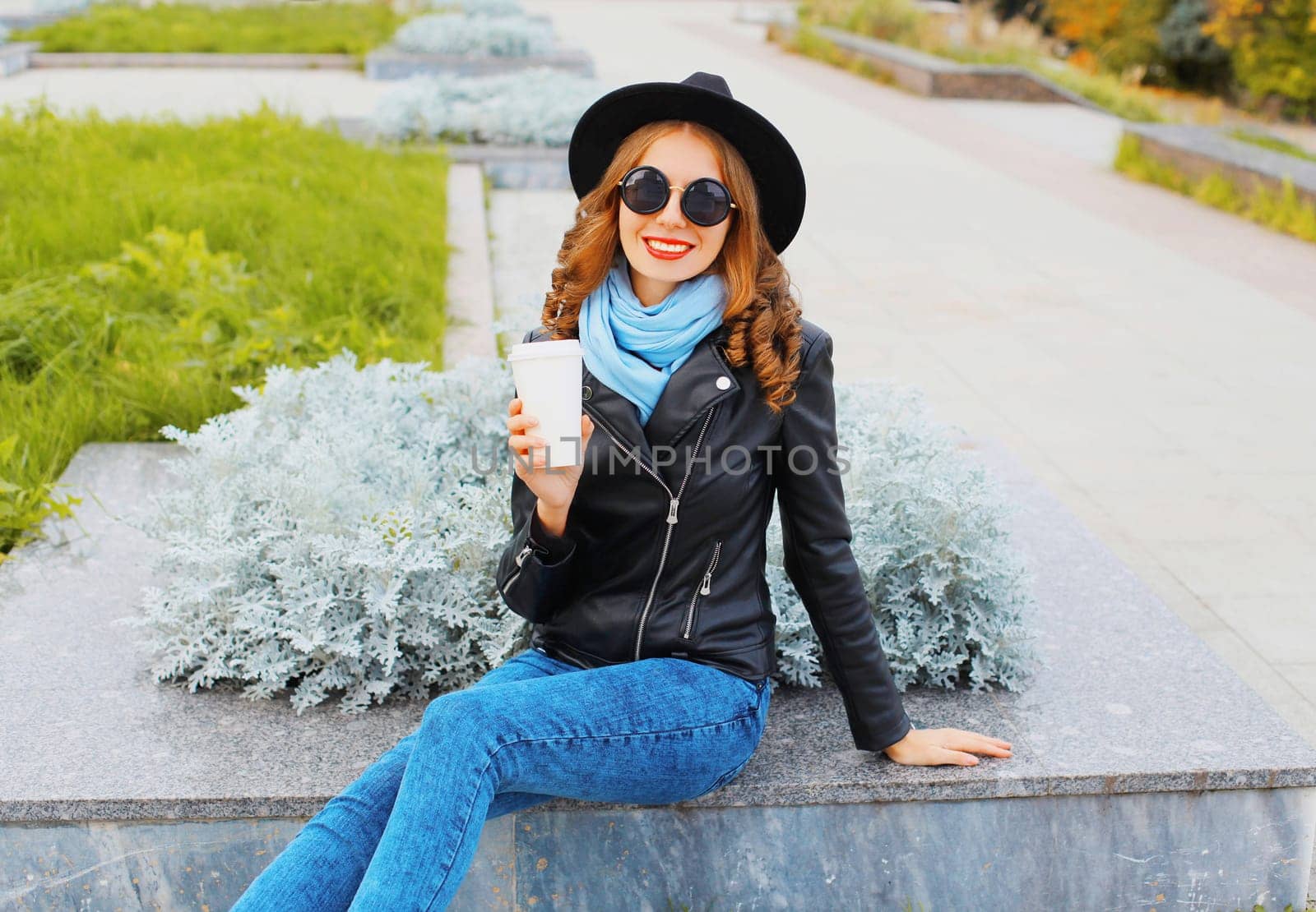 Fashion cool smiling girl with coffee cup wearing a black rock jacket, hat in city park