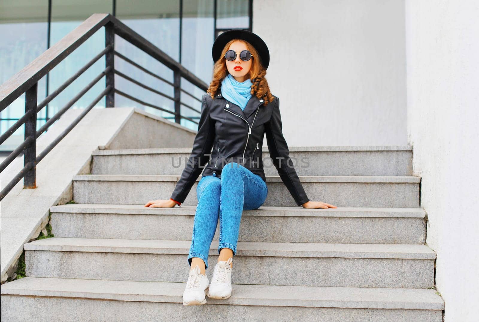 Fashion portrait pretty young woman wearing a black rock jacket and hat sitting in city