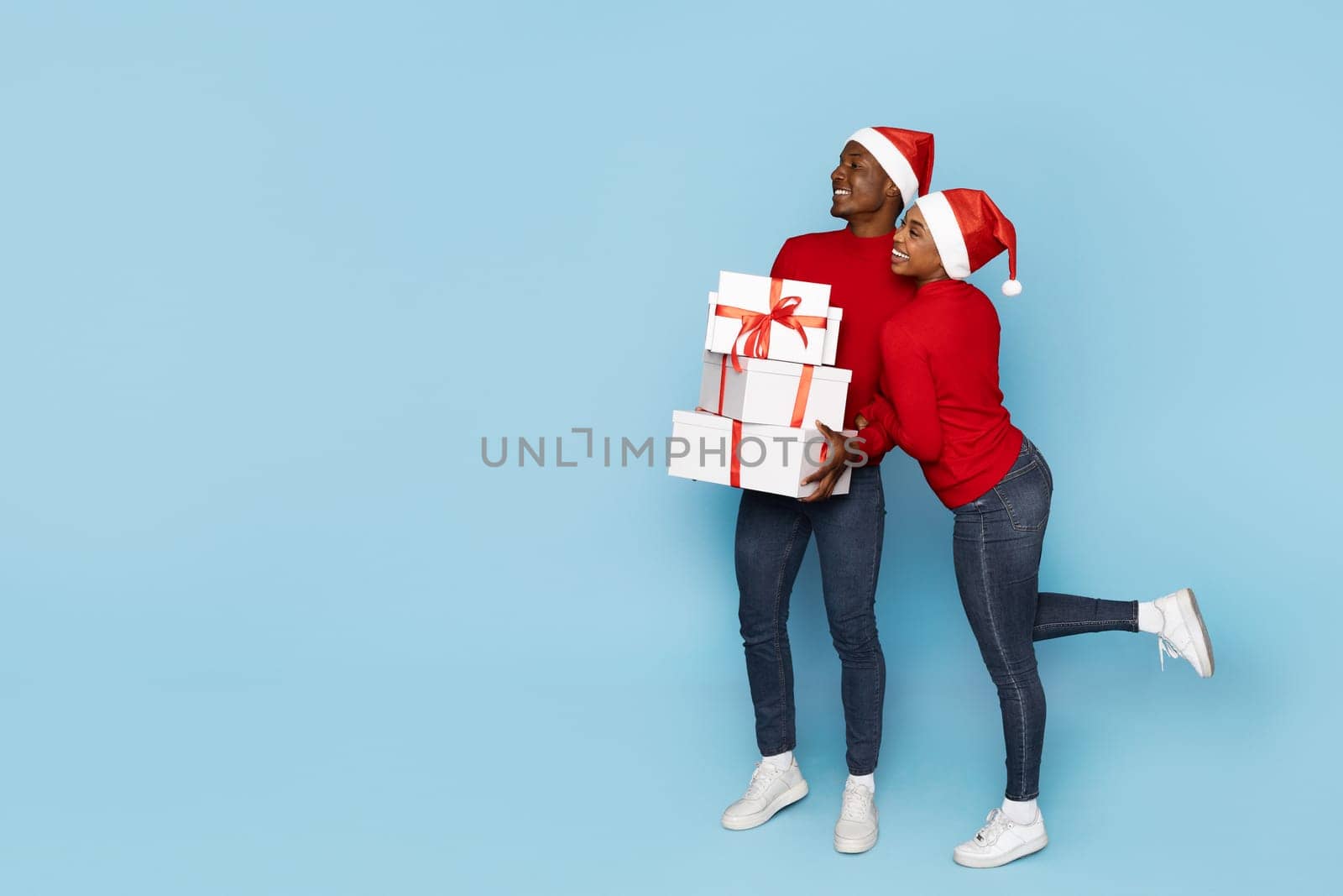 A cheerful couple in matching red sweaters and Santa hats embraces while holding beautifully wrapped gifts against a light blue background, embodying holiday spirit and joy, copy space
