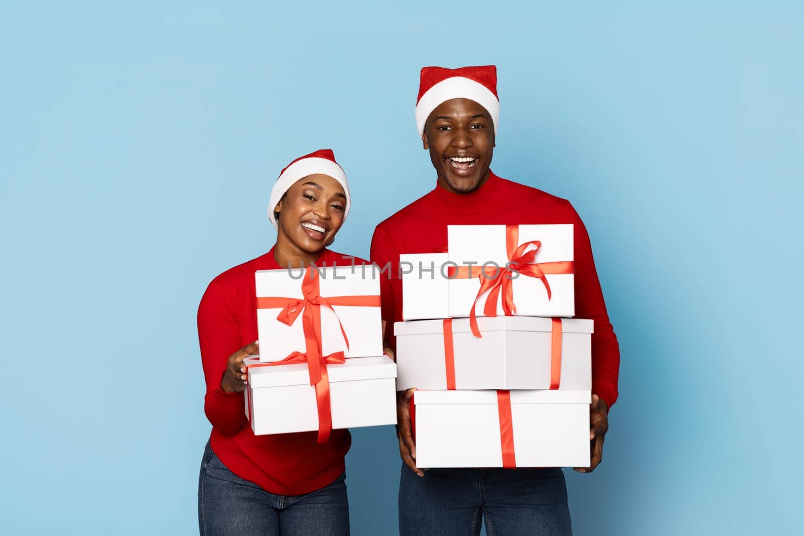 A couple dressed in bright red sweaters and Santa hats happily presents wrapped Christmas gifts with red ribbons, celebrating the festive holiday season together.
