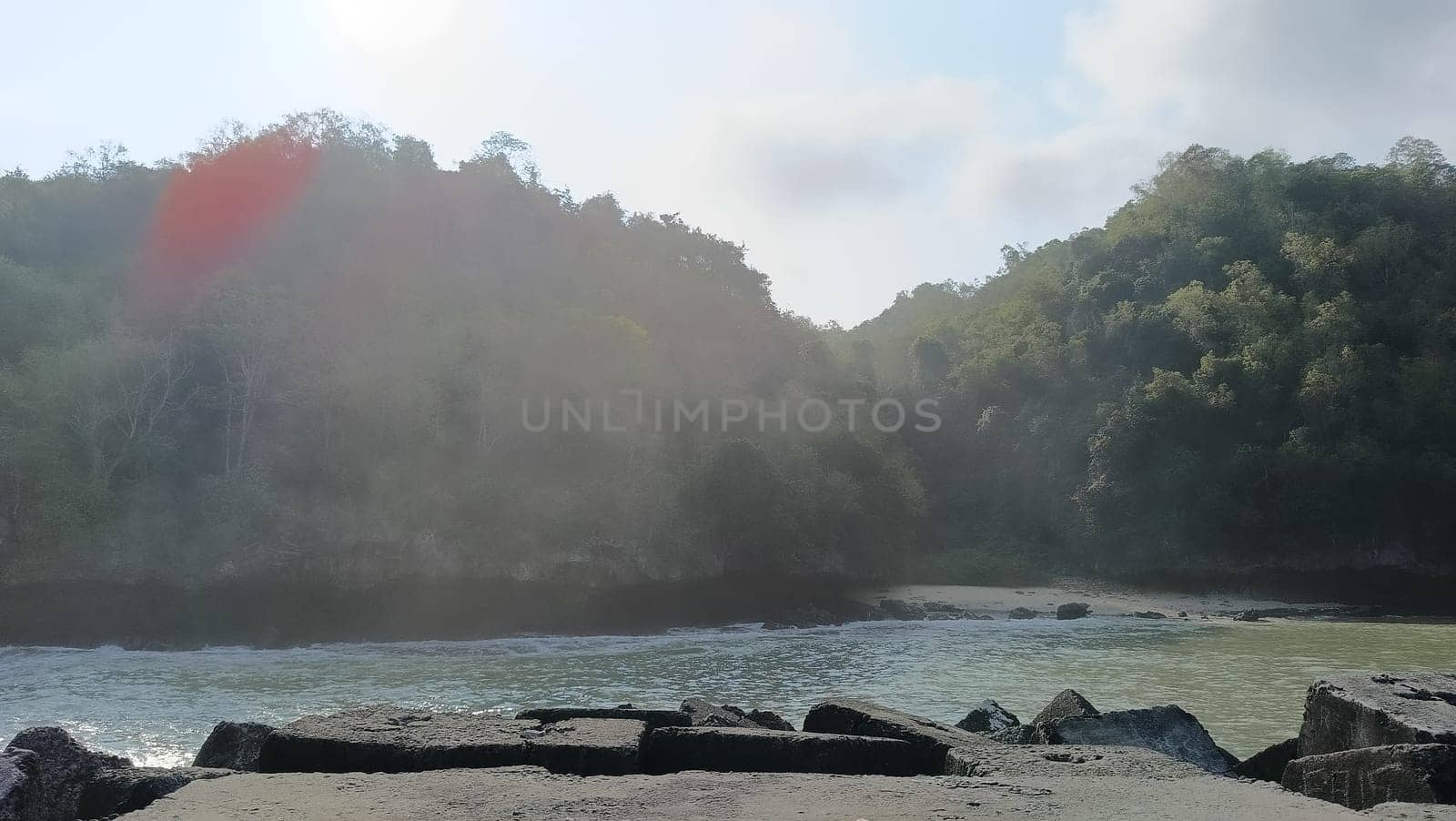 A rocky shoreline with a large rock formation in the background. The water is calm and the sky is blue