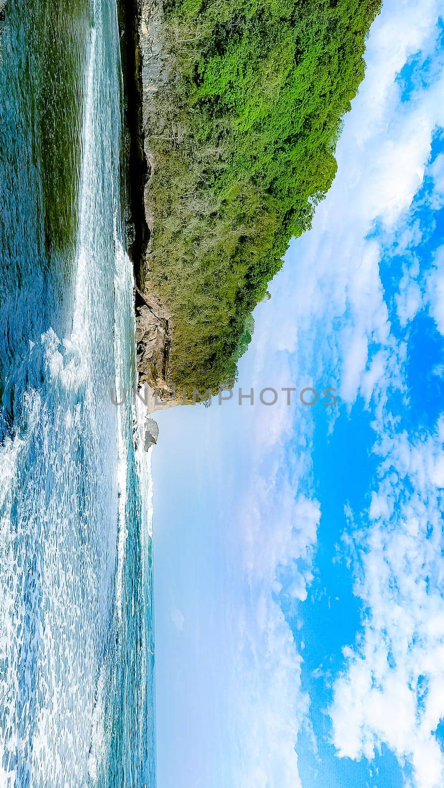A rocky shoreline with a large rock formation in the background. The water is calm and the sky is blue
