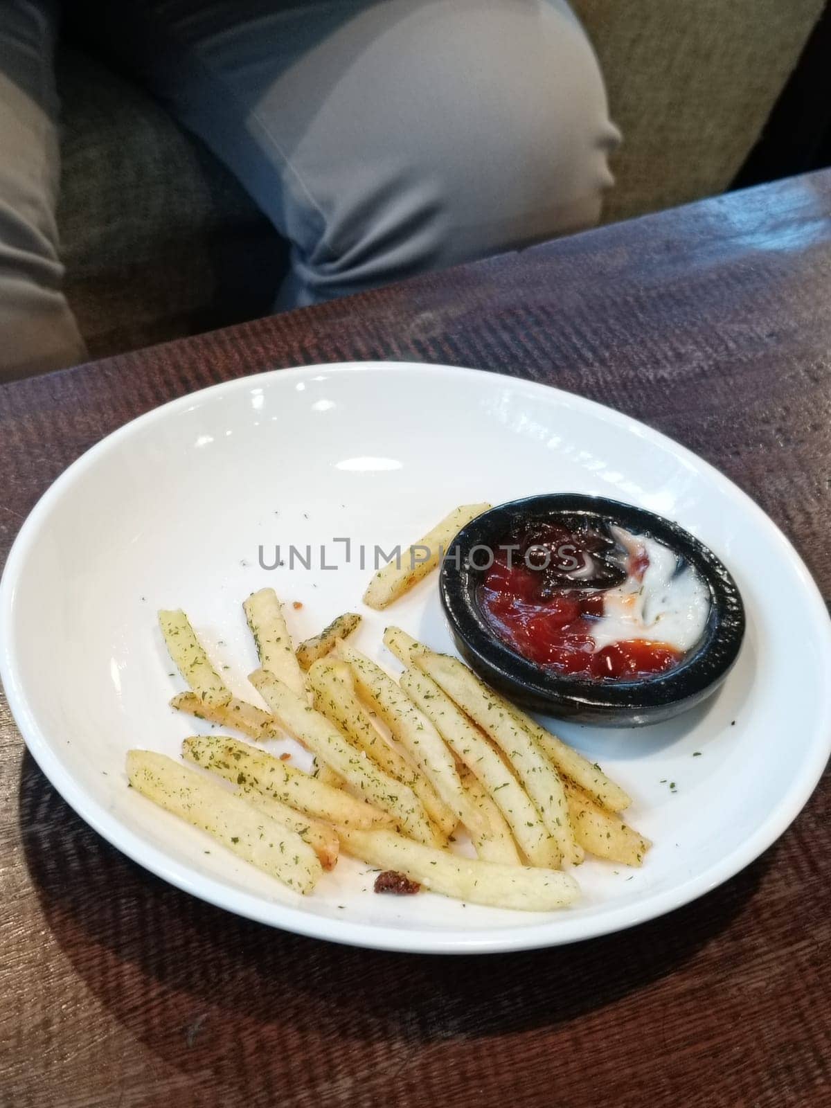 A bowl of savory French fries seasoned with herbs, accompanied by ketchup and mayonnaise, served on a rustic wooden table. Perfect for snack time or a casual dining setting.