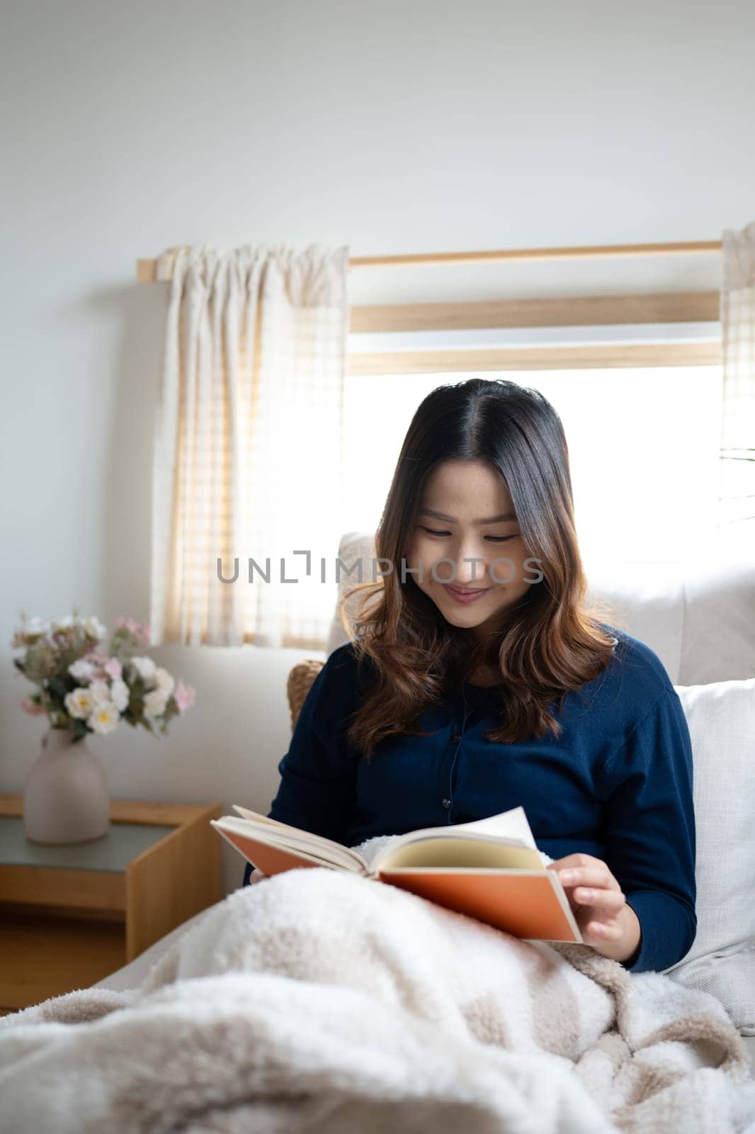 Smiling young woman reading a book by the Window, wrapped in soft blanket.