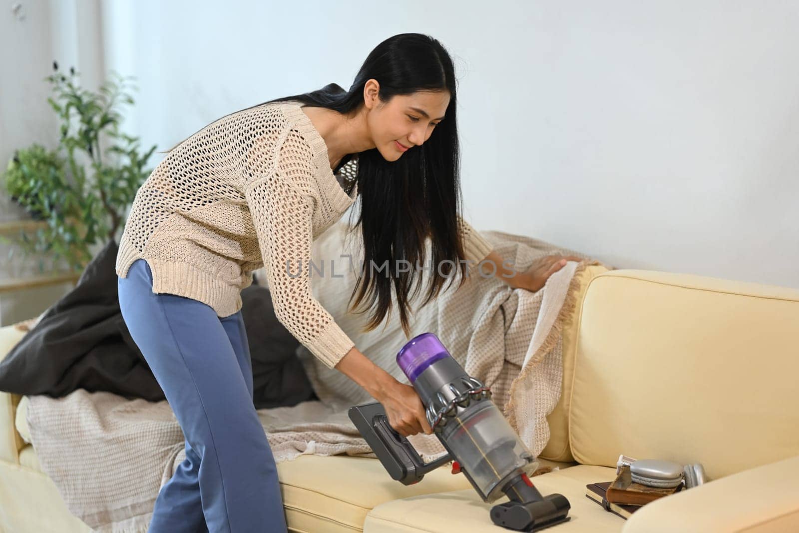 Young woman cleaning her living room sofa with a cordless vacuum.