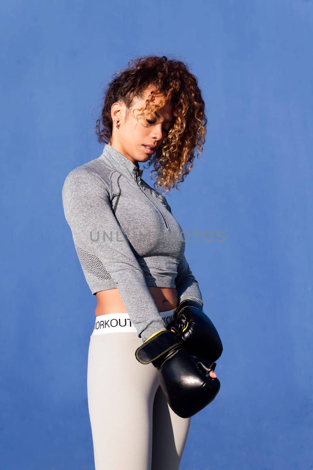 determined adult female boxer putting on boxing gloves during workout on blue background, concept of strength and healthy lifestyle