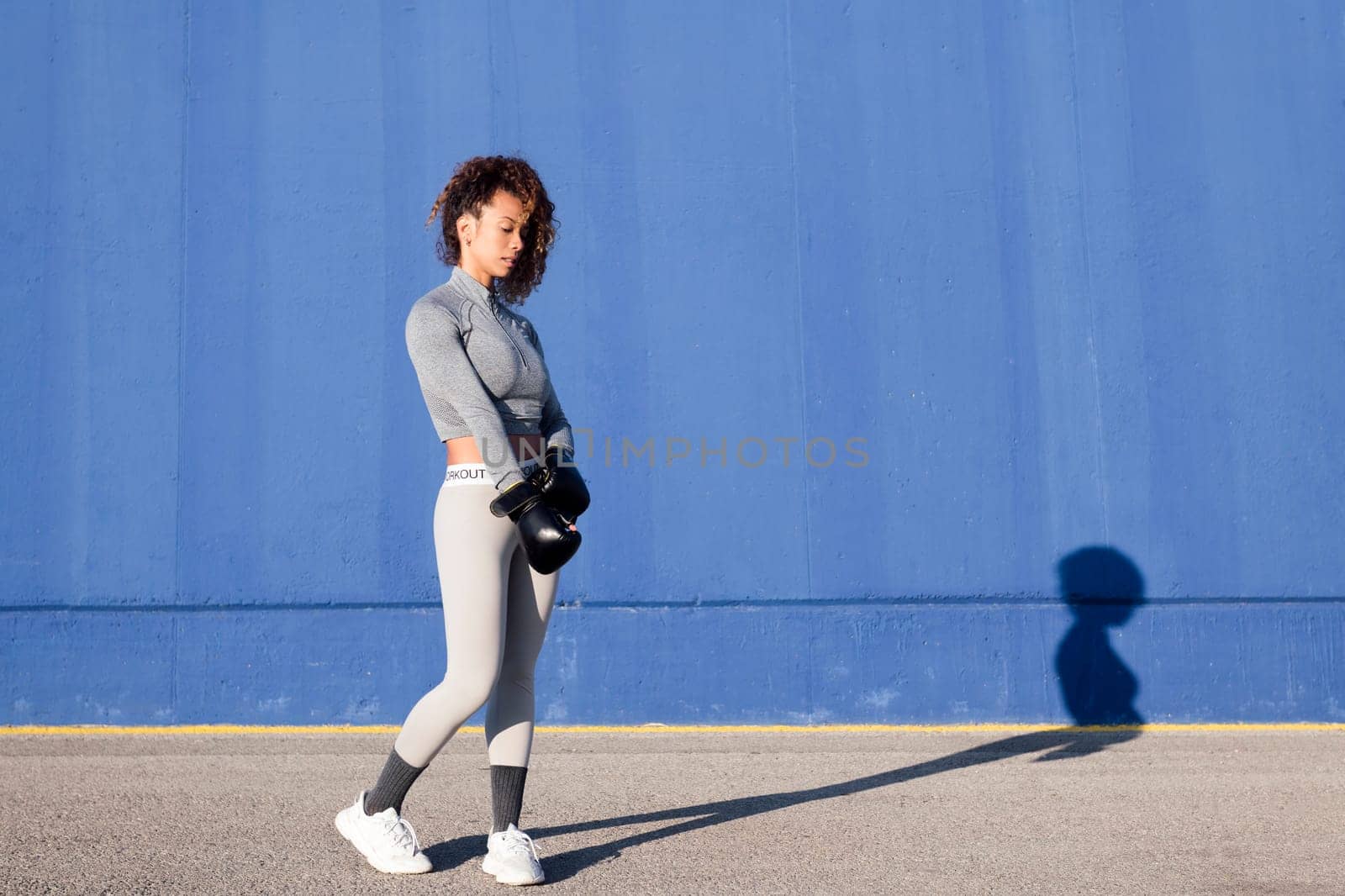 determined adult female boxer putting on boxing gloves during workout on blue background, concept of strength and healthy lifestyle, copy space for text