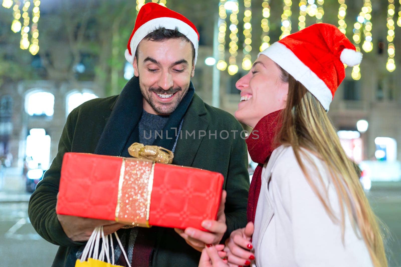 A happy couple wearing Santa hats exchange gifts outdoors, enjoying the festive holiday spirit. The man holds a beautifully wrapped present as they share a laugh, surrounded by glowing lights.