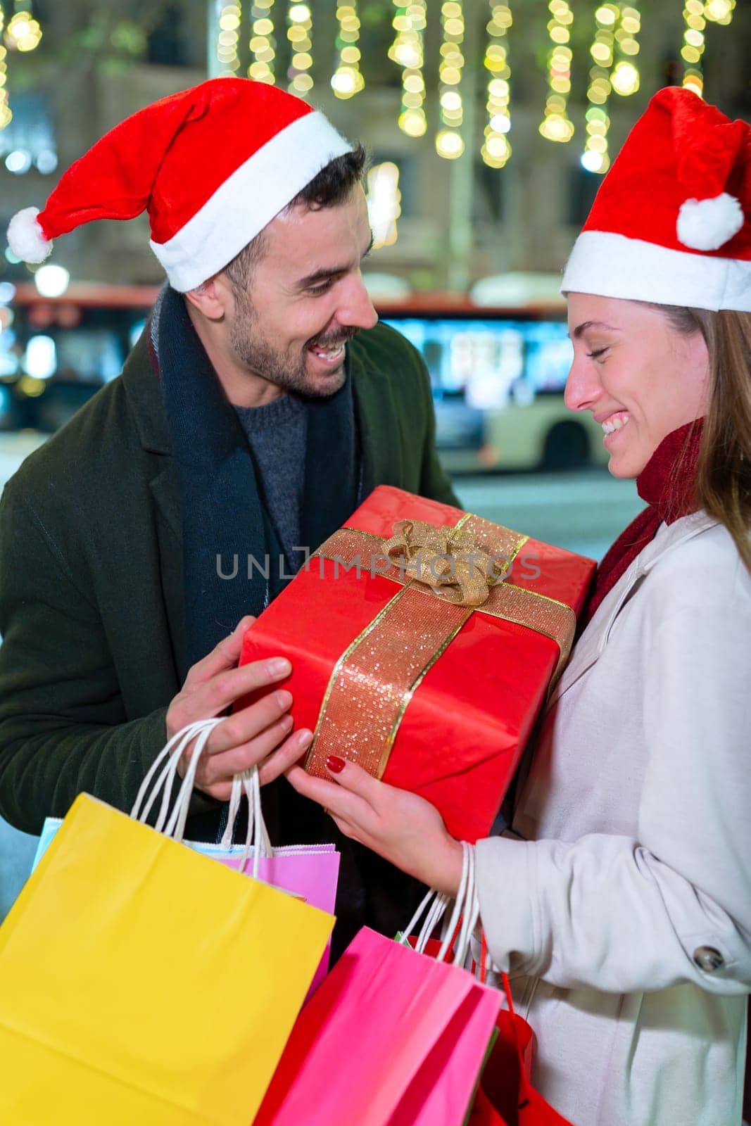 A happy couple wearing Santa hats exchange gifts outdoors, enjoying the festive holiday spirit. The man holds a beautifully wrapped present as they share a laugh, surrounded by glowing lights.Vertical