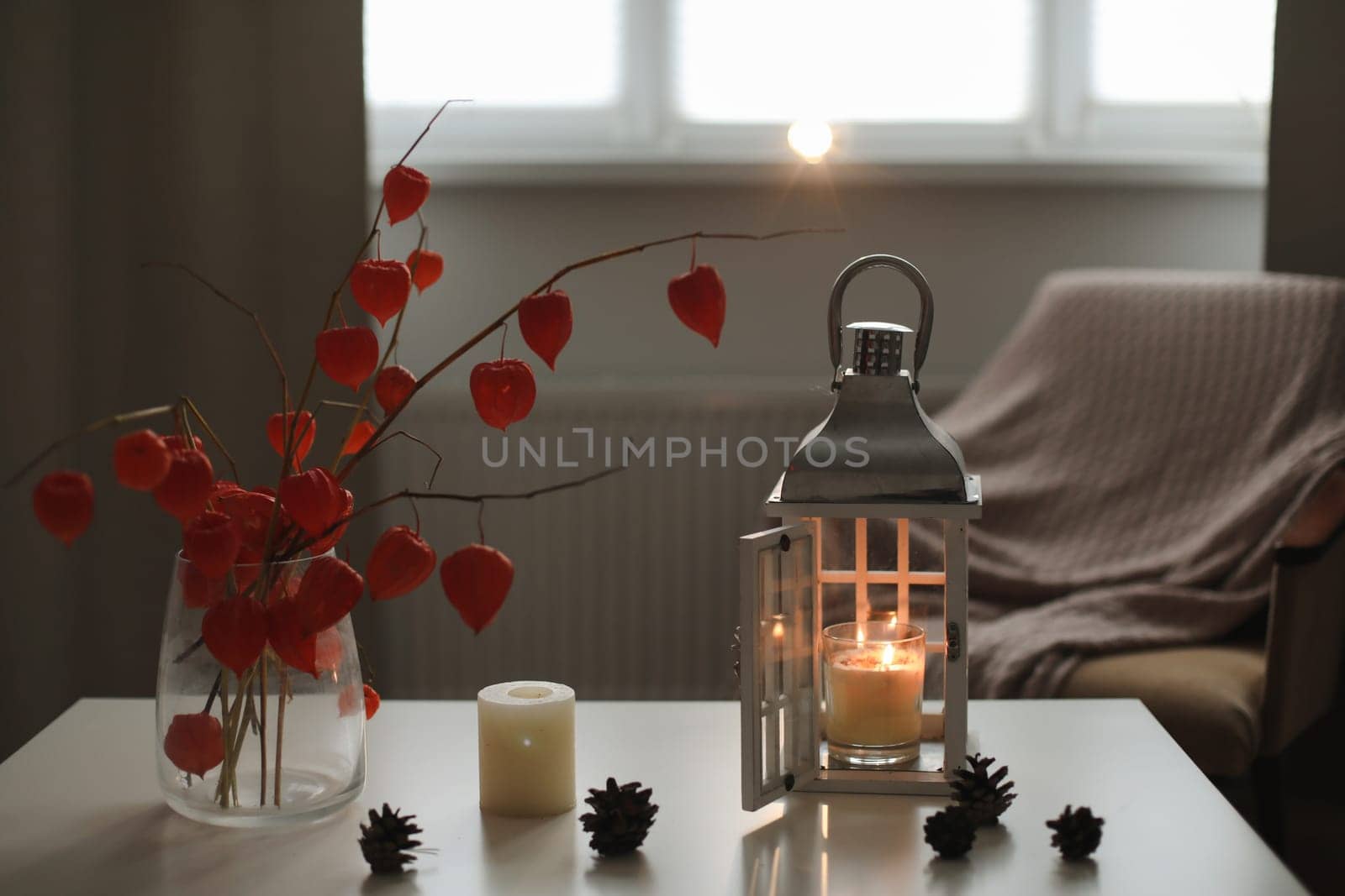 autumn still life with candles, flowers, twigs of red physalis in a vase in a cozy home interior.