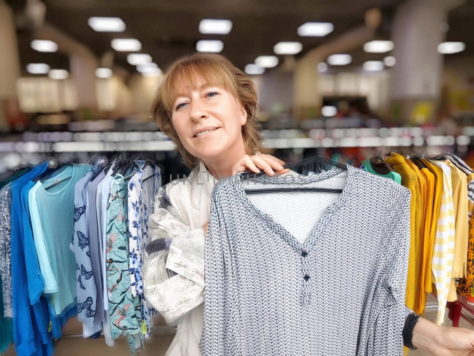 A middle aged woman enjoys shopping in a retail store, excitedly holding a clothing item while browsing colorful selections.