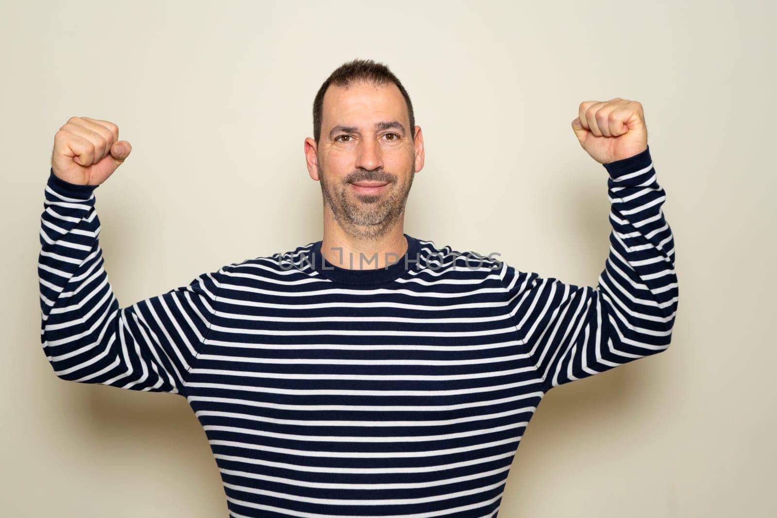 Bearded hispanic man in his 40s wearing a striped sweater flexing his biceps while smiling proudly without realizing the deplorable physical condition he is in, isolated over beige background
