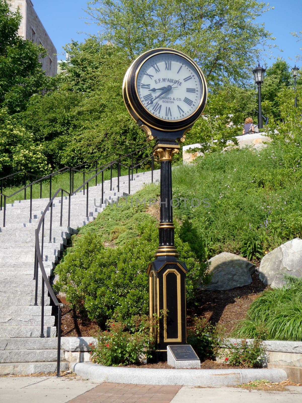 Boston - May 30, 2010: A beautifully crafted clock tower sits beside stone steps, surrounded by lush greenery in the park, creating a picturesque atmosphere on a sunny day.