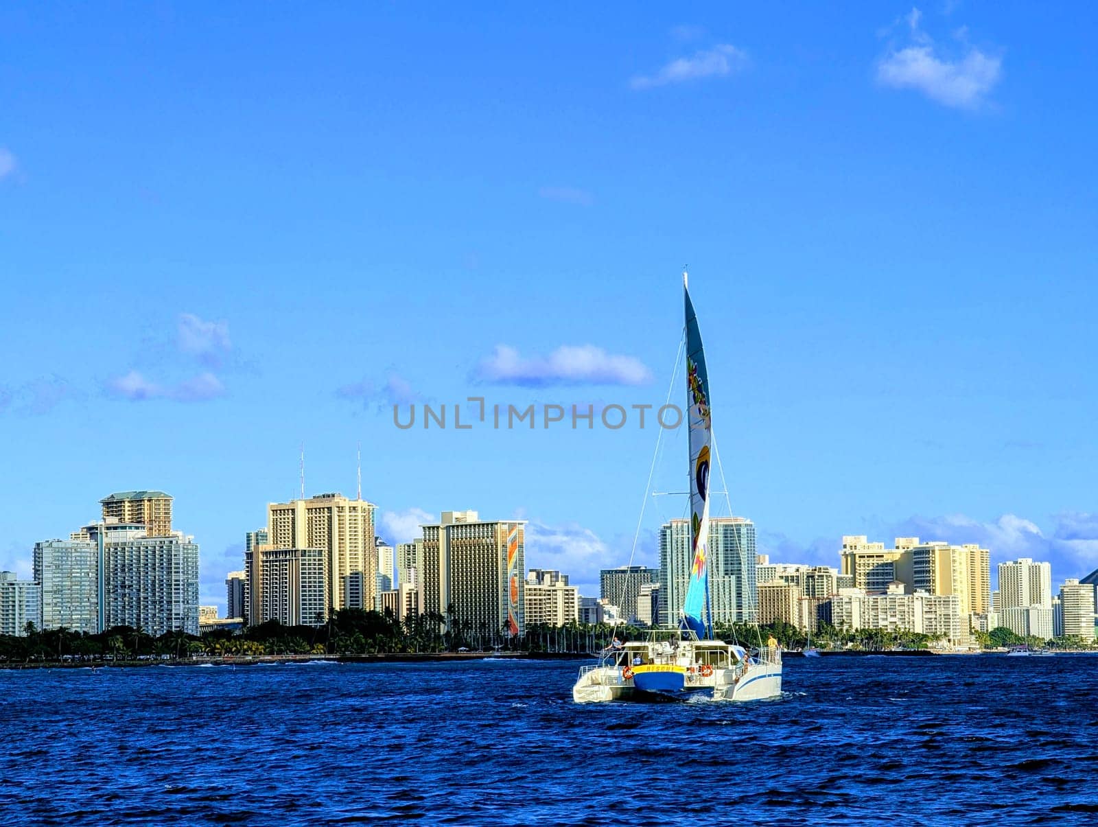 Honolulu - October 13, 2024: A sailboat navigates the shimmering waters, with the bustling Honolulu skyline providing a stunning backdrop against a bright blue sky.