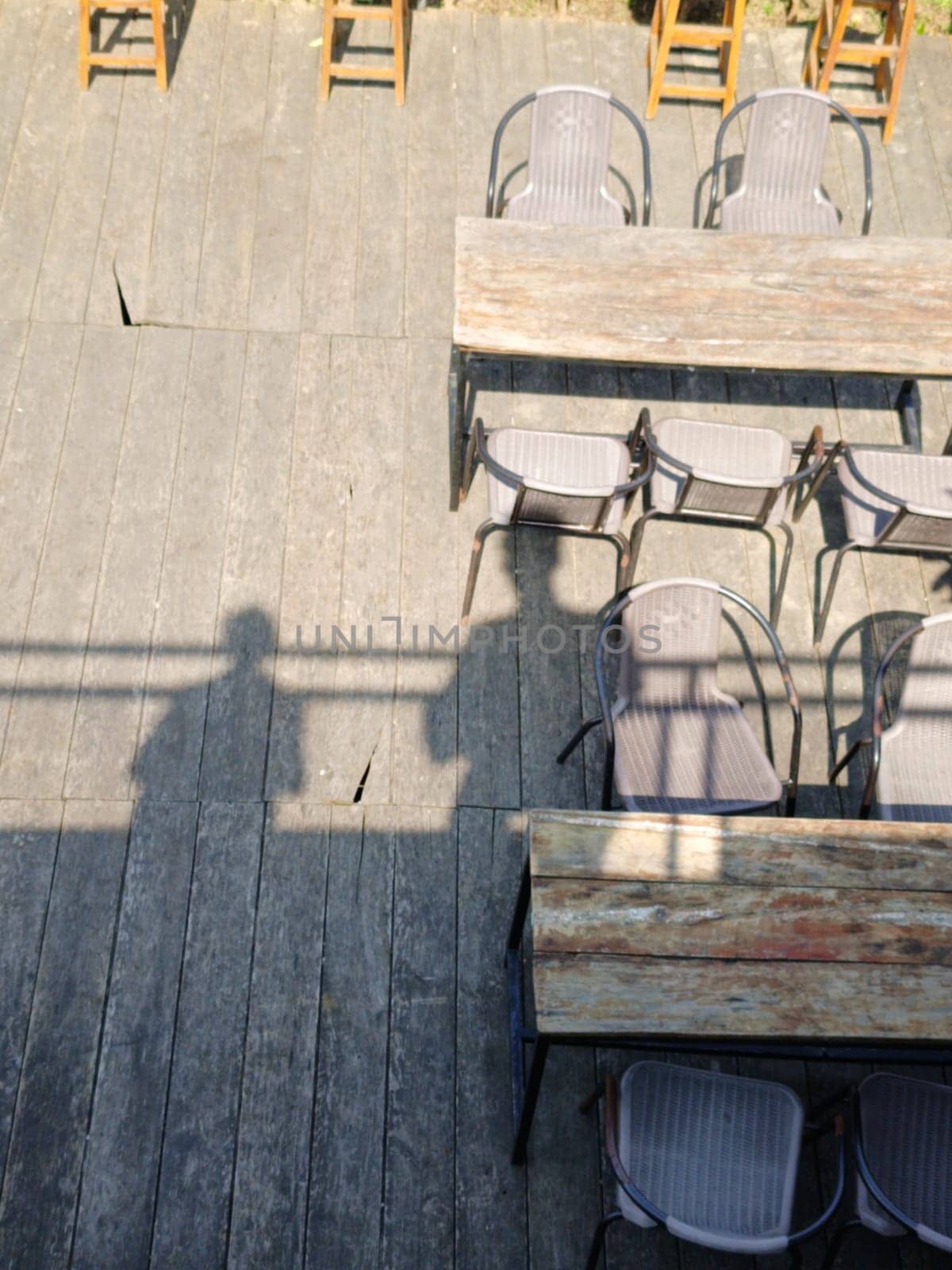 Top view of empty rustic wooden tables with chairs on an outdoor wooden deck, creating a serene and inviting atmosphere. There is also shadows of people from a rooftop