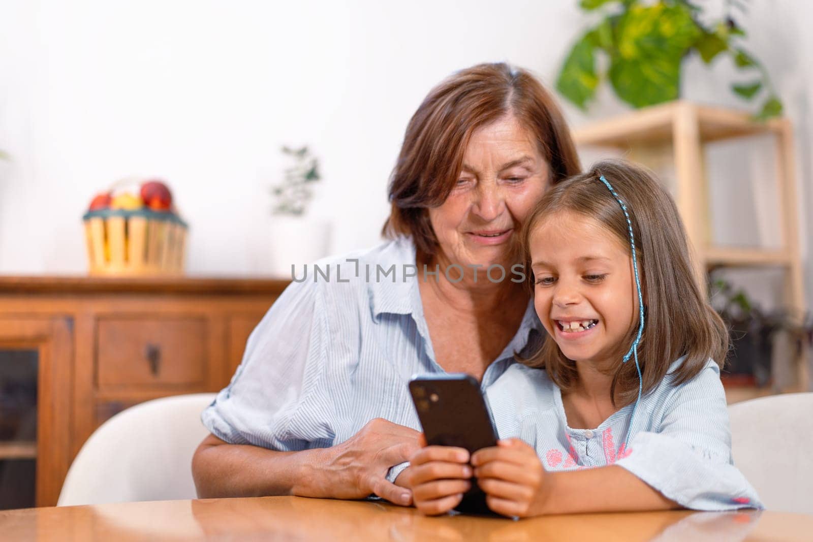 Happy granddaughter using smartphone with grandmother on table at home. Their smiling faces are full of content as they share and learn from the gadget