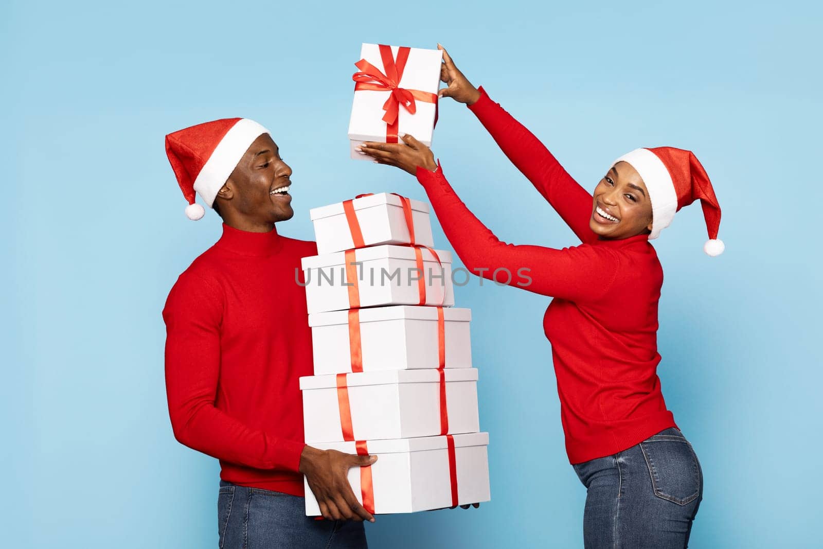 A black couple wearing red sweaters and Santa hats joyfully presents a tall stack of gift boxes wrapped in red ribbons. Their cheerful expressions enhance the festive spirit of the moment.