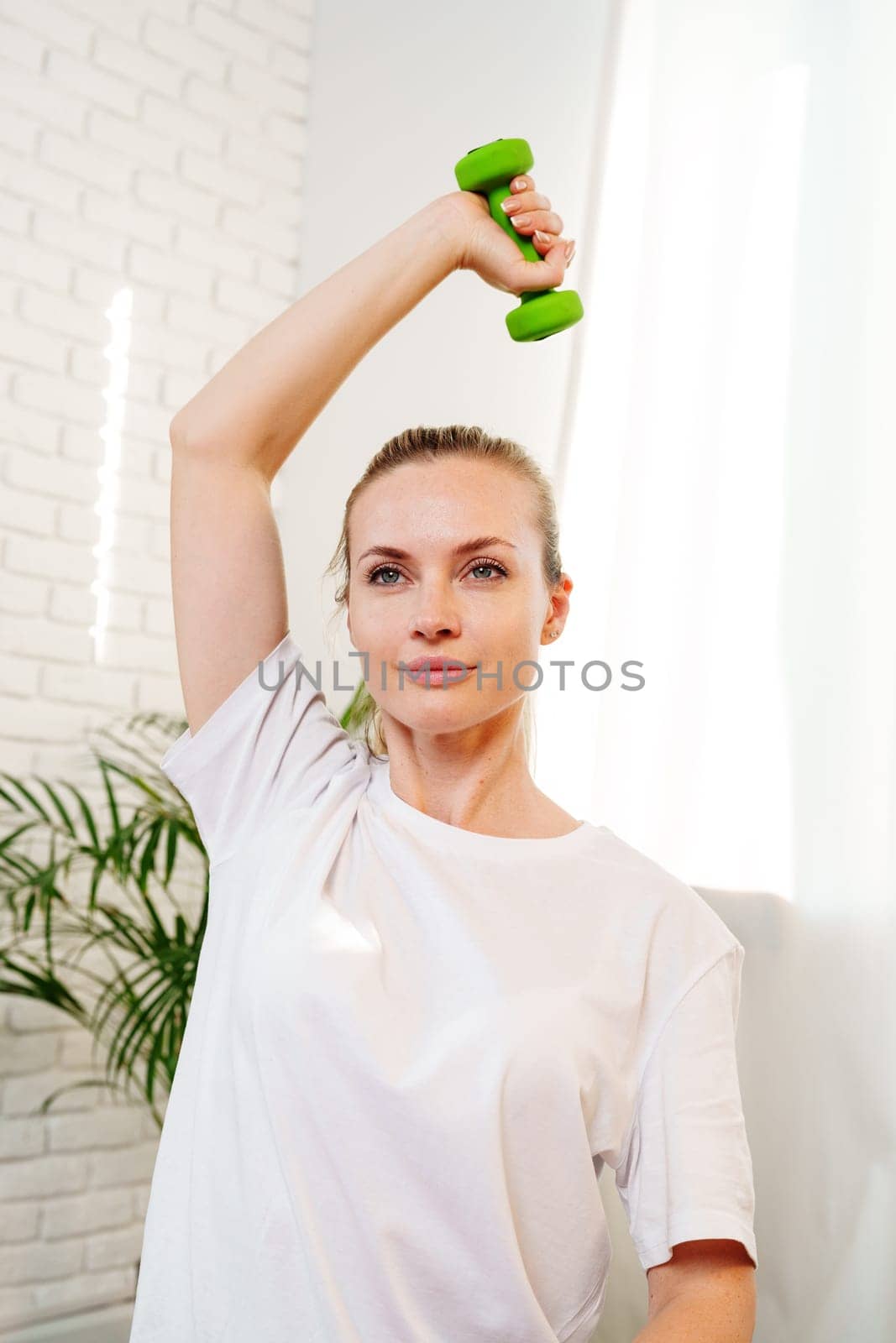 A woman exercises with green dumbbells at home in a bright room filled with light and plants