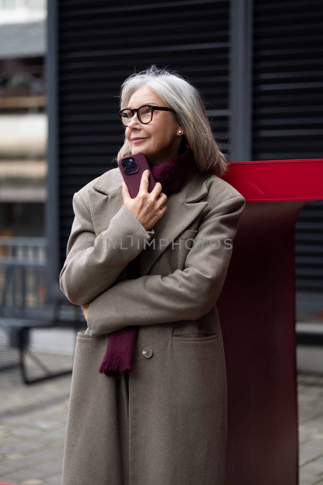 A senior businesswoman stands with a relaxed demeanor outside, dressed warmly in a coat and scarf. She holds a smartphone to her chest, enjoying the cool air.