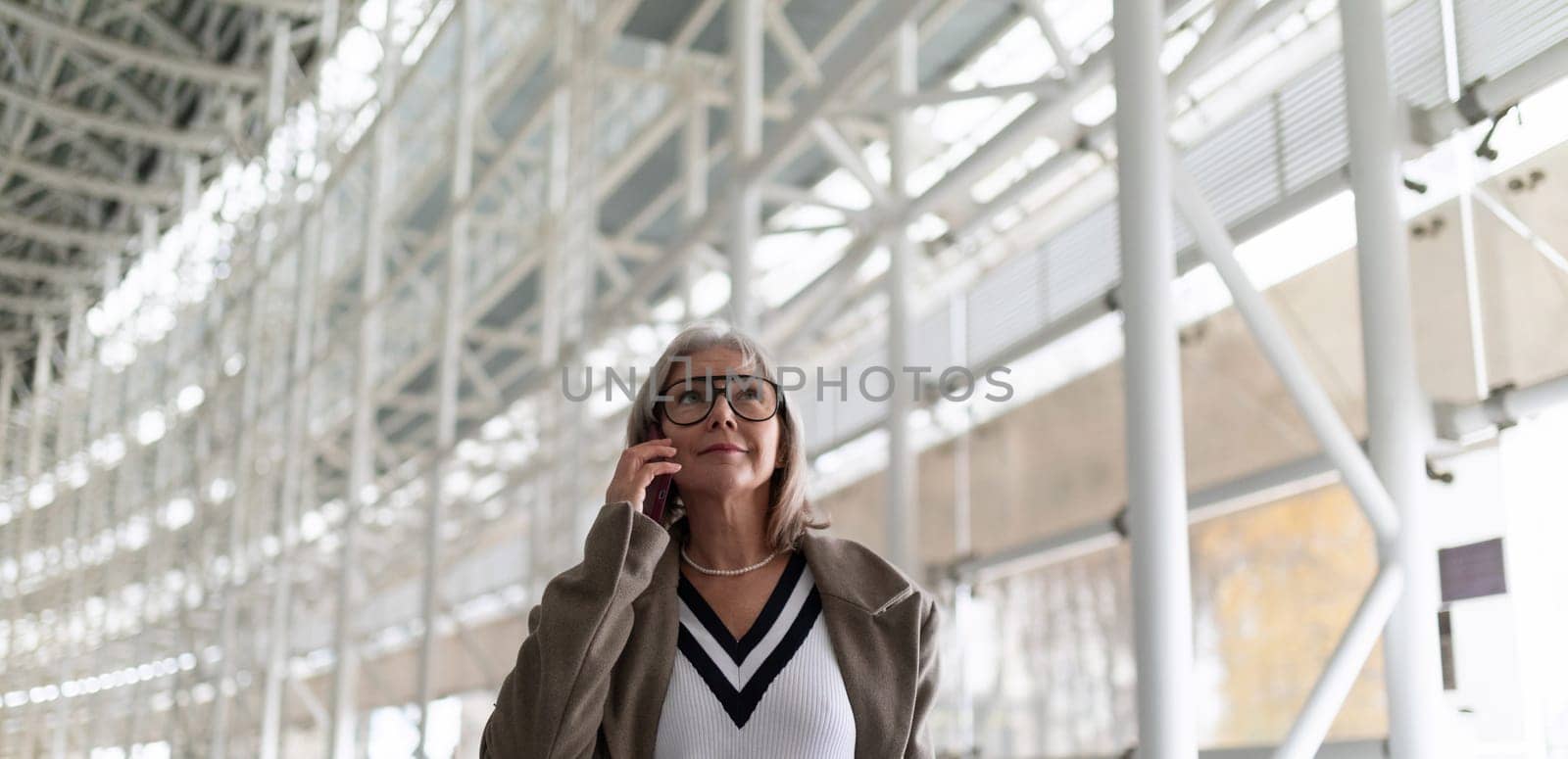 A middle-aged businesswoman with glasses is engaged in a phone conversation while strolling through a bright and spacious airport terminal. Her confident demeanor suggests professionalism and purpose.