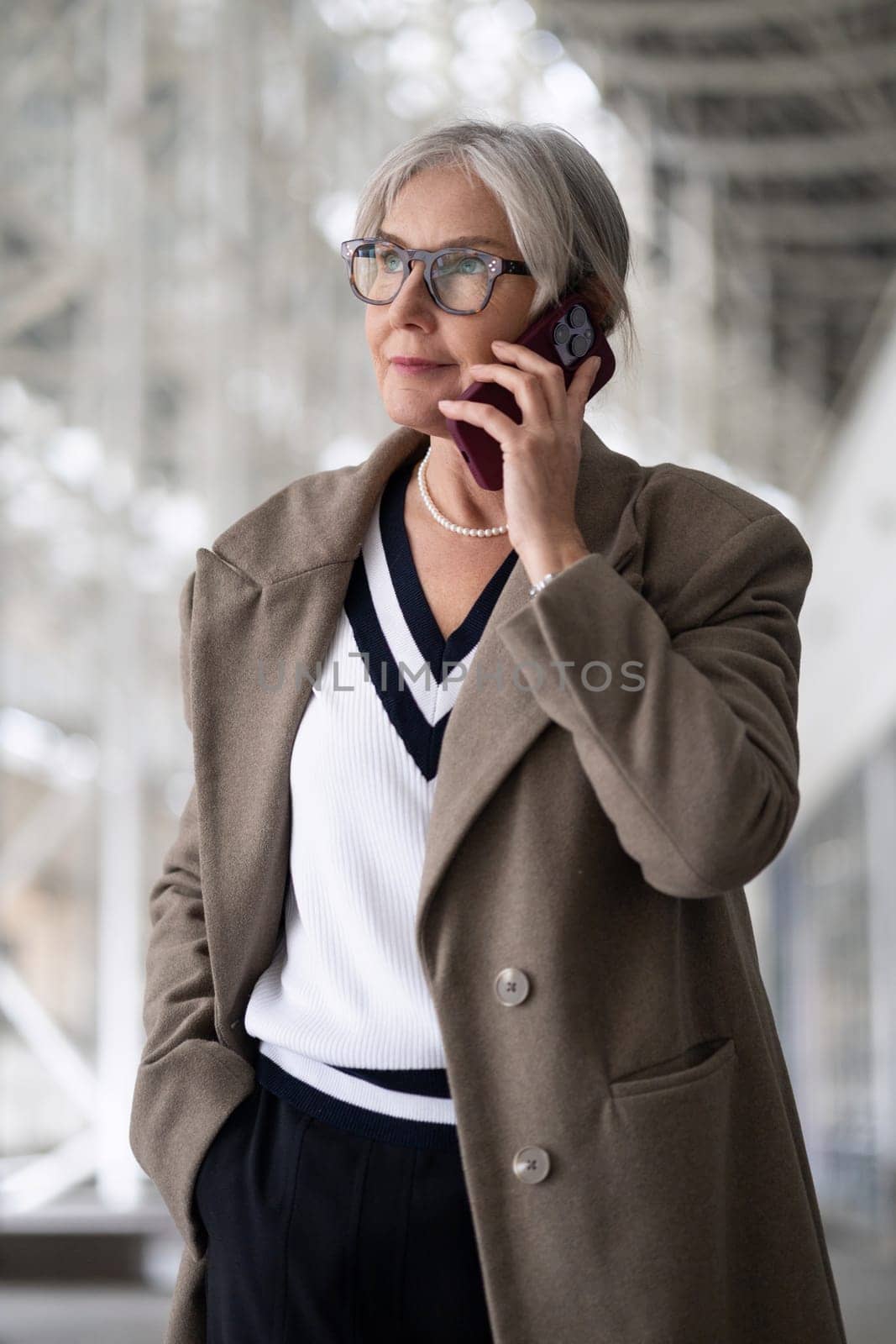 A senior businesswoman stands outside a contemporary office building, speaking thoughtfully on her mobile phone, dressed in professional attire, exuding confidence.