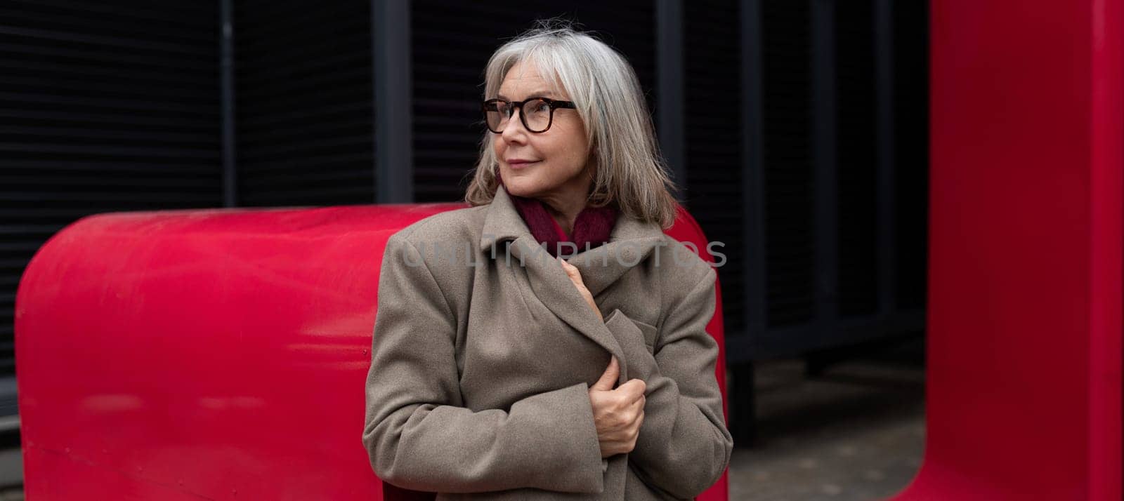 An elderly businesswoman poses gracefully in a fashionable beige coat, showcasing her confidence while surrounded by a colorful red backdrop in an urban environment.