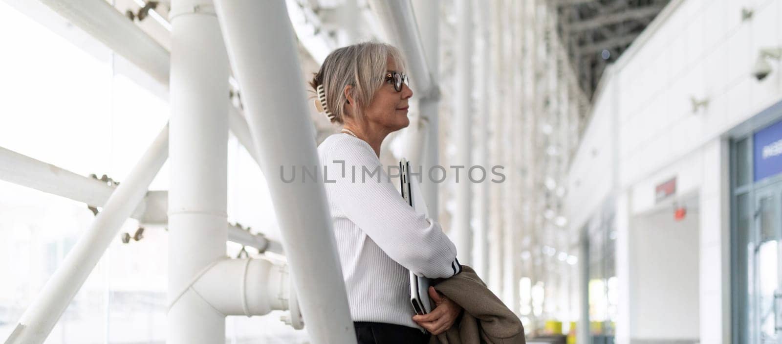 A middle-aged businesswoman stands confidently in a sleek office, reviewing important documents. She appears focused and engaged in a professional setting.