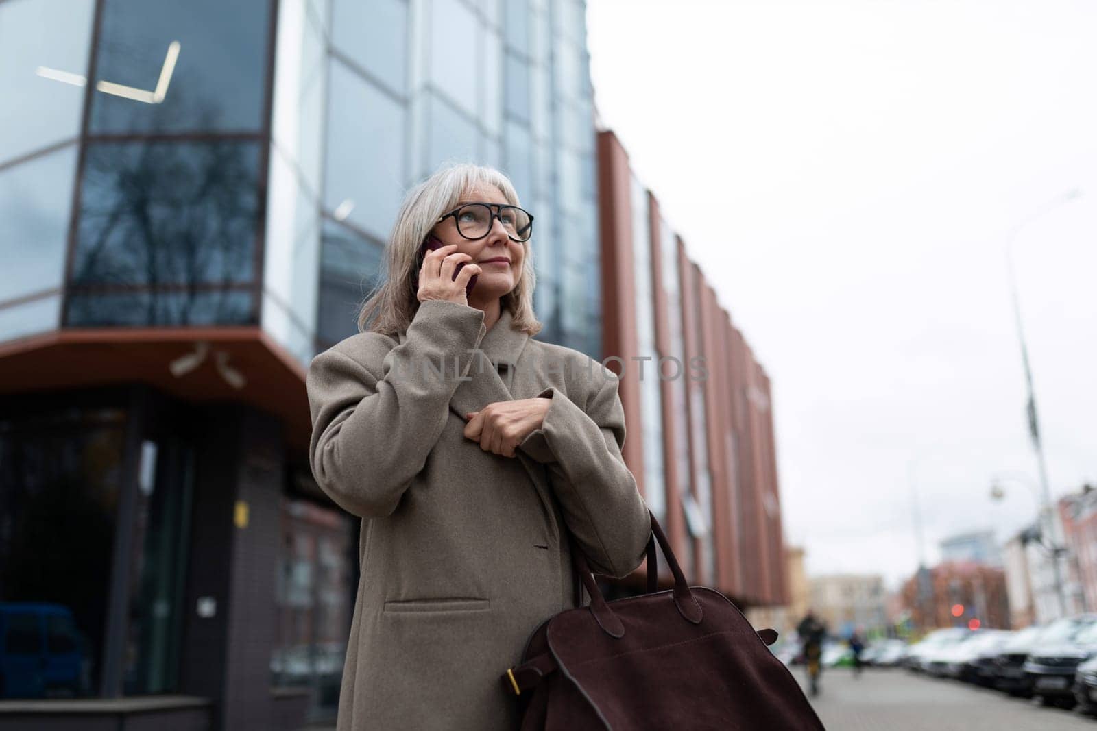 A middle-aged businesswoman dressed in a stylish coat stands on the street, engaged in a phone conversation while holding her handbag. The urban setting features modern buildings.