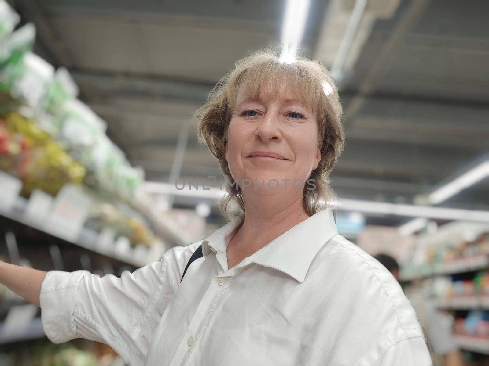 A middle aged woman smiles while selecting fresh produce in a brightly lit supermarket, enjoying her shopping experience.