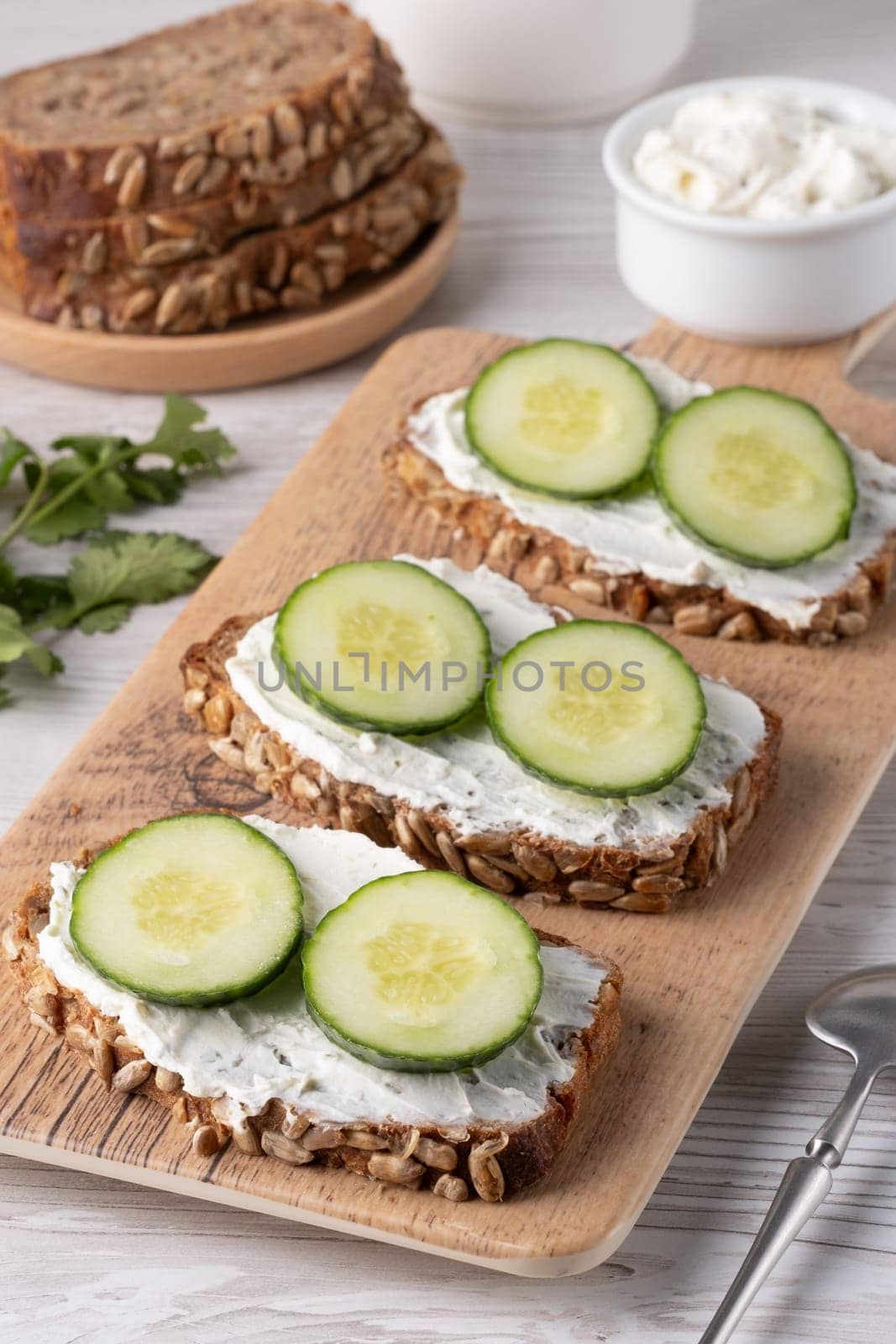 Three slices of nutritious whole grain bread are spread with creamy cheese and topped with fresh cucumber rounds, arranged on a wooden board for a light snack.