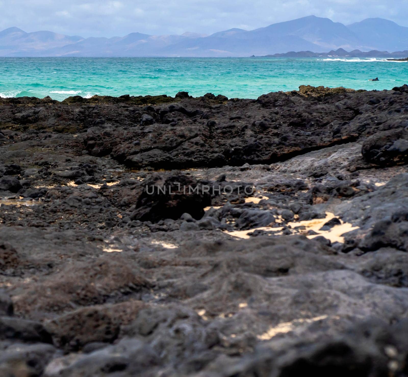 Beach with black volcanic sand and stones on the coast of the Atlantic ocean. A beautiful light blue clean water and mountains on the horizon, a picturesque shore by the sea without people.