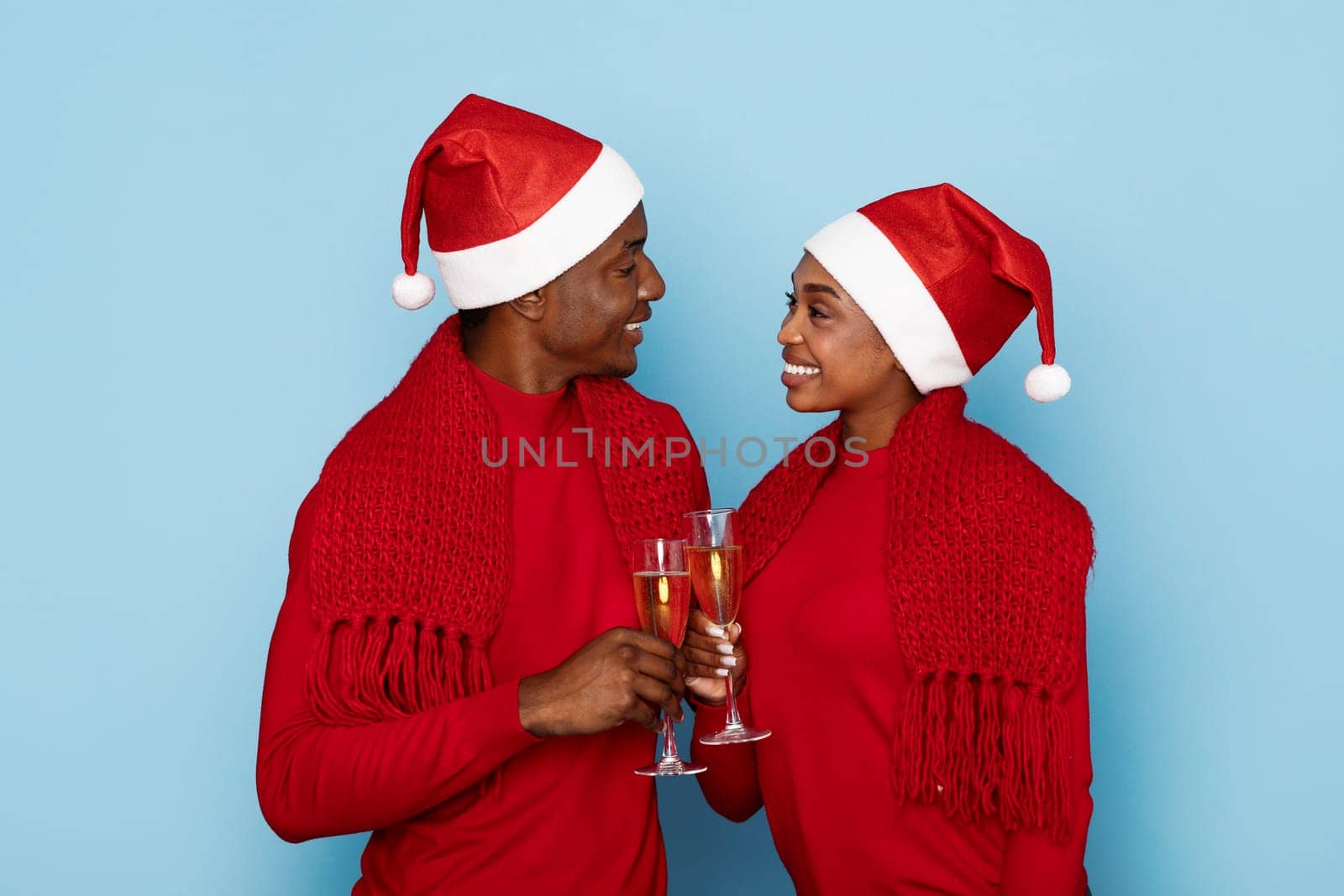 A joyful couple in red outfits, adorned with Santa hats, clinks champagne glasses together in a festive celebration. Their bright smiles radiate holiday cheer against a light blue background.