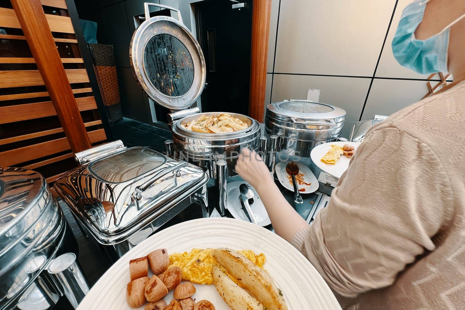 A lady is at the hotel buffet, loading up her plate with food from a food warming machine. From the viewpoint of the individual, food is being artfully arranged on a plate.
