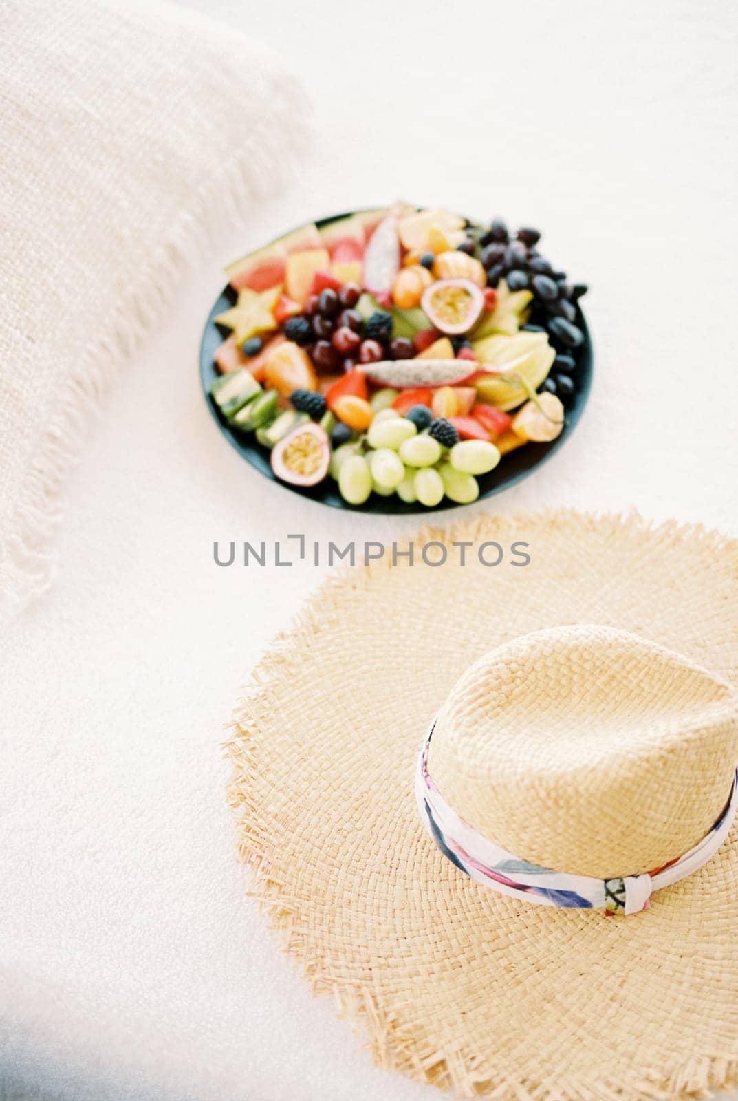 Plate of chopped fruit stands on a white sheet on the bed next to a straw hat. High quality photo